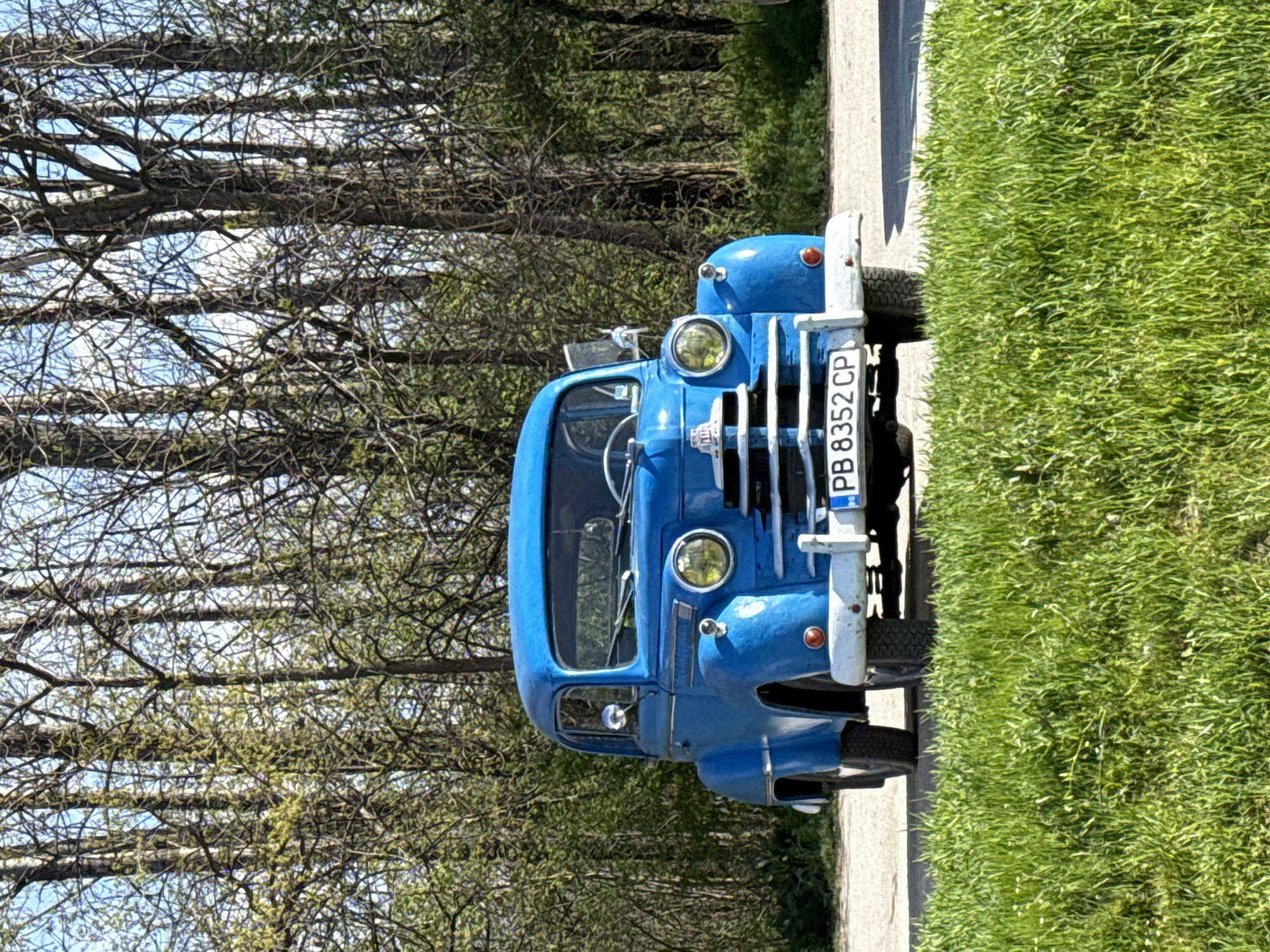 A blue vintage car with a license plate reading "PB 8352 CP" is parked on a paved road alongside a grassy area. The car is positioned facing the camera, with a background of trees, some bare and some with sparse foliage. The location is likely in Plovdiv, Bulgaria, based on the license plate format.