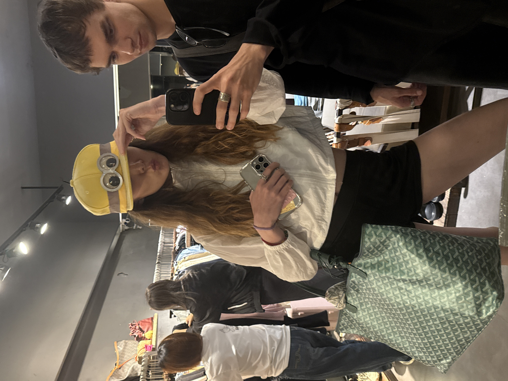 In Xuhui District, China, a young woman wearing a yellow hat with googly eyes and a white jacket is taking a selfie with a young man in a black t-shirt. The woman is holding a phone in her left hand and is making a pouting face. She is also wearing a short black skirt and carrying a green tote bag. The man is wearing glasses and has his arm around the woman. In the background, there are racks of clothing and bags, suggesting that they are in a store.