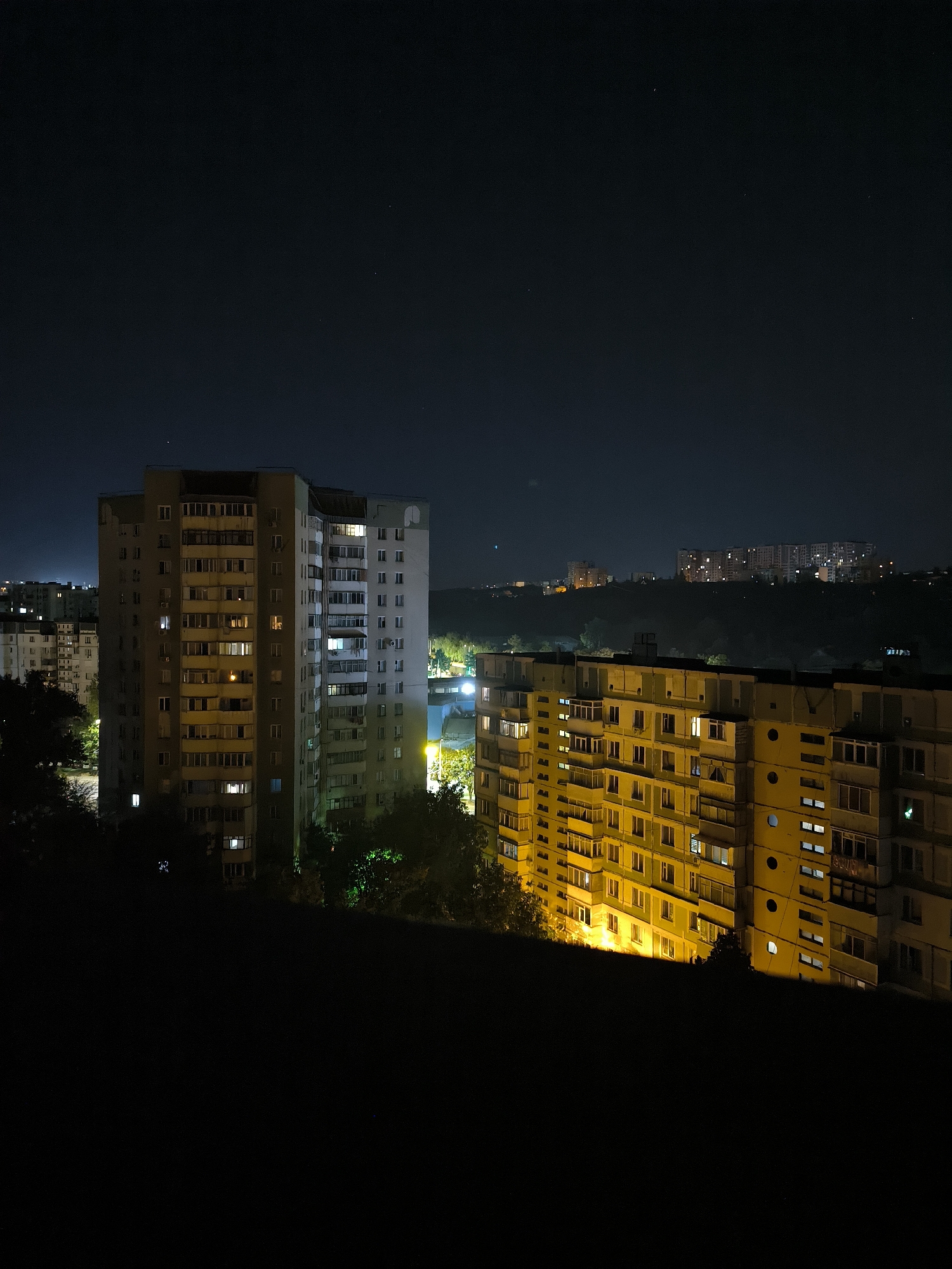 The image shows a night view of a residential area in Chișinău, Moldova. Two multi-story apartment buildings are prominent in the foreground, illuminated by lights from within the apartments and streetlights. The buildings appear to be Soviet-era panel buildings, characterized by their uniform structure and balconies. Trees and vegetation are visible between and in front of the buildings, adding a natural element to the urban landscape. In the background, other apartment buildings and structures are visible, stretching across a hilly terrain under a dark night sky dotted with a few stars. The overall scene depicts a typical residential neighborhood at night, with signs of human habitation through the lit windows.