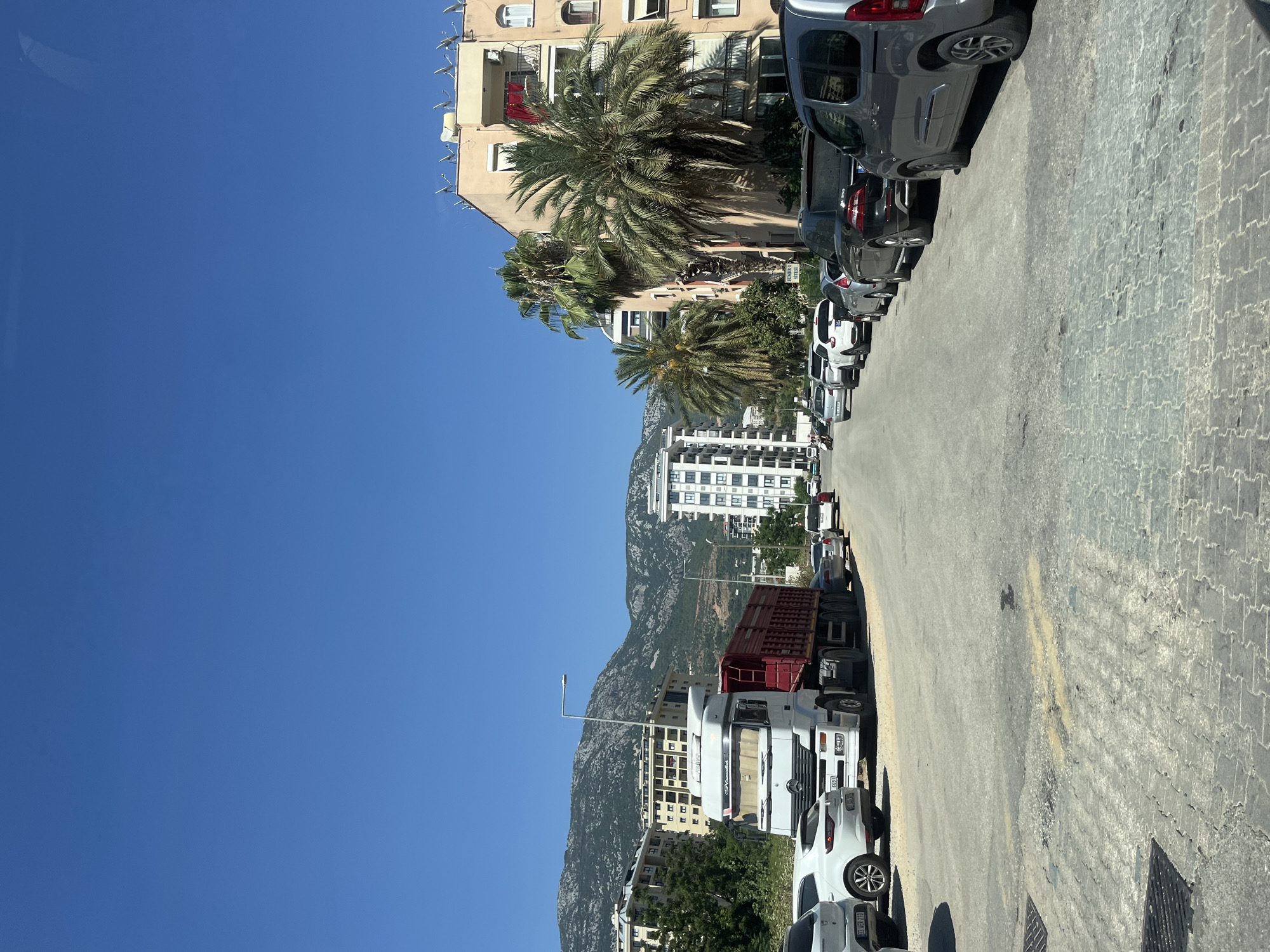 A view of a residential street in Alanya, Turkey, on a clear sunny day. The street is lined with parked cars on both sides. In the foreground, the road surface is paved with interlocking stones that are worn and cracked in places. There are metal grates embedded in the pavement on the left.
On the left side of the street, a large white truck with a red trailer is parked. Further down the street, several cars are parked, including a white sedan and a gray minivan. Behind the parked cars, there are multi-story apartment buildings with balconies and satellite dishes visible on the rooftops. Green trees and palm trees are interspersed between the buildings.
In the background, a large mountain range with rocky slopes and sparse vegetation rises behind the city. The sky is a clear, bright blue. The overall impression is a typical urban street scene in a coastal city with a warm climate.