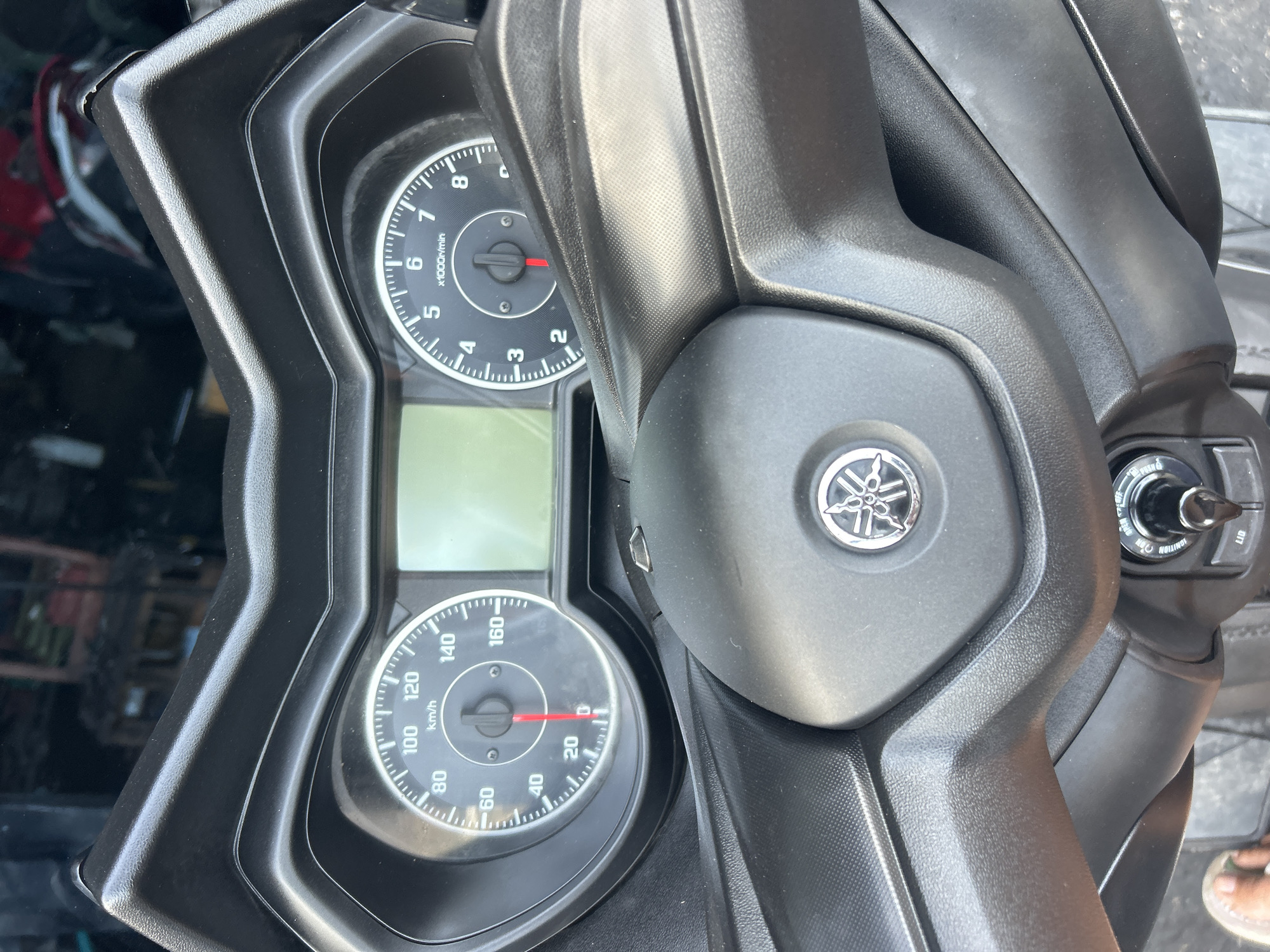 The image shows a close-up view of a motorcycle's dashboard, specifically the speedometer and tachometer. The speedometer is on the left, marked in km/h, with the needle pointing slightly above 0. The tachometer is on the right, marked in x1000r/min, with the needle pointing between 0 and 1. A digital display is located between the two gauges. The dashboard is made of black plastic, with a Yamaha logo prominently displayed on the steering column. A key ignition is visible at the bottom of the image. The background is blurred, showing a dark, indistinct environment, possibly an indoor storage area. There are no people clearly visible in the image. This scene is not specifically identifiable as being in Ko Samui District, Thailand.