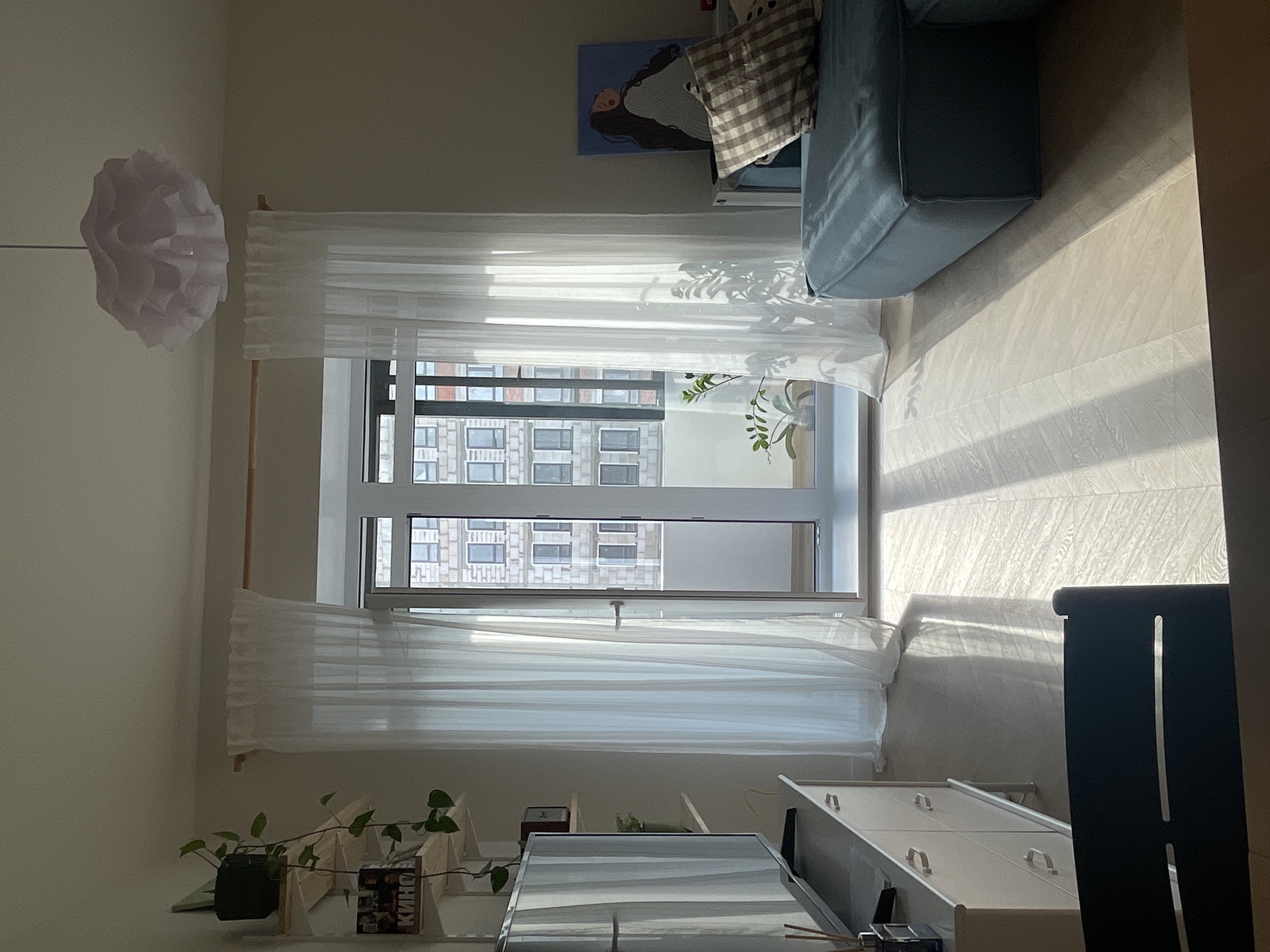 The interior of a room is depicted with a large window allowing natural light to flood in. Sheer white curtains hang on either side of the window. Outside the window is a view of a building with multiple floors and windows, suggestive of an urban setting. A blue sofa with a plaid pillow is situated near the window. To the right of the window, a white dresser is visible. A large window that provides natural light into the interior of the room and plants can be seen near the window. The room is decorated with white, soft furnishings. The location is Izhevsk, Russia, evident from the book titled "КИНО" on the shelf, which is written in Russian.
