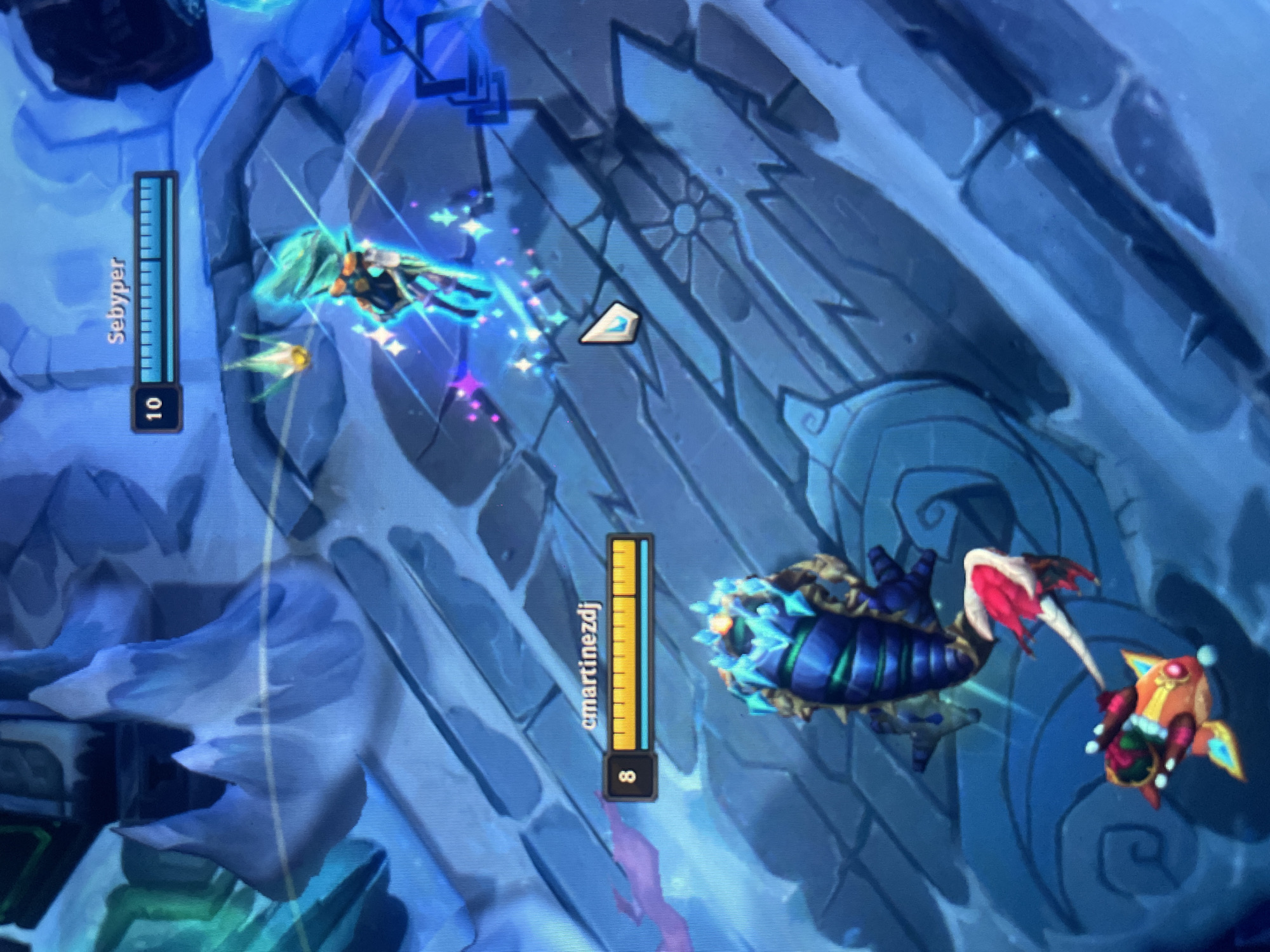 This image depicts a scene from a video game, likely a MOBA given the user interface elements. Two players' characters are engaged in action. On the left, a character with a light blue and white ethereal design, surrounded by sparkling particles, is in motion. A health bar above this character displays the name "Sebyper" and a level indicator of "10". To the right, a blue and dark purple, bug-like creature with glowing blue crystalline protrusions is moving. This creature is wielding a staff adorned with red and white fabric and a golden headpiece. A health bar above this creature shows the name "cmartinezdi" and a level indicator of "8". The background is a stylized, icy, and mountainous landscape with geometric structures and subtle blue and purple lighting, suggesting a fantasy or sci-fi setting. The overall atmosphere appears dynamic and action-oriented. There are no discernible cues about the time of day or weather. The location context of Huixquilucan de Degollado, Mexico, is external information and not visually present in the image.