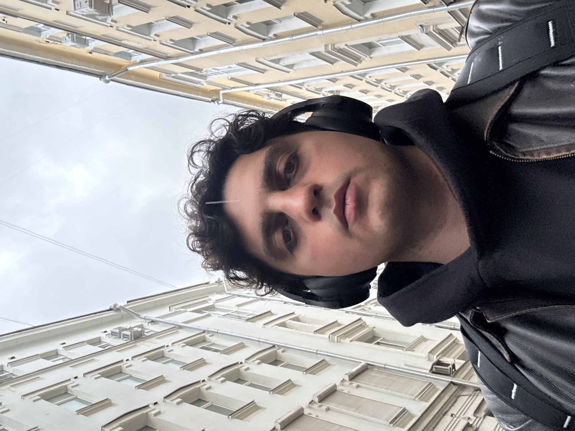 A low-angle self-portrait of a man with curly dark hair wearing black headphones, a black hooded sweatshirt, and a black leather jacket with backpack straps. He is looking down at the camera. The background consists of multi-story buildings with windows and architectural details, suggesting an urban setting. Based on architectural style and details visible in the background buildings, the image was captured in Moscow, Russia. The sky appears overcast.