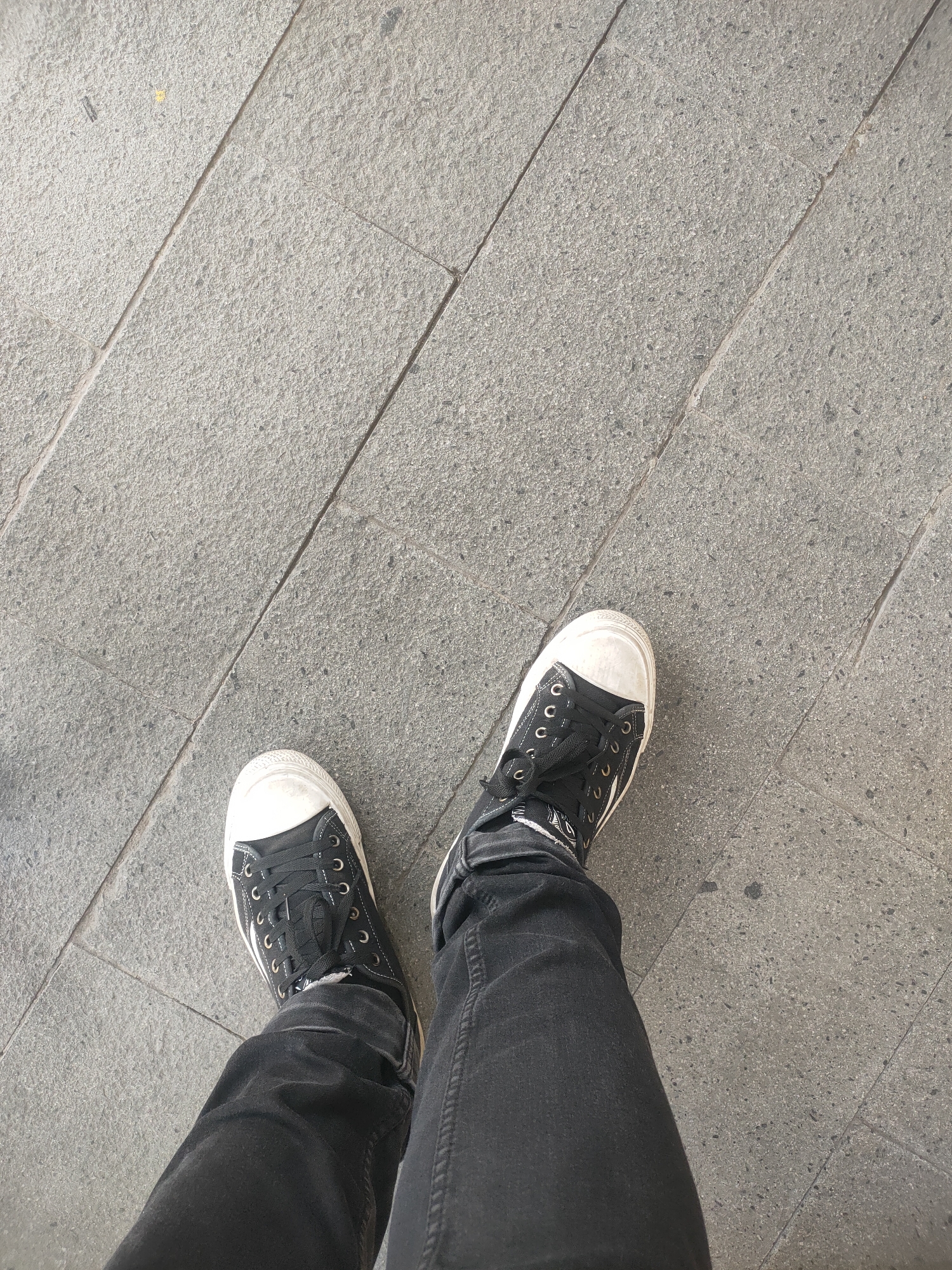 The image shows a low-angle perspective of a person's legs and feet standing on a tiled sidewalk. The person is wearing black jeans and black high-top sneakers with white laces and white toe caps. The tiles are large, rectangular, and gray with visible grout lines. The scene appears to be outdoors in a city environment. The specific location is identified as Sooko, Indonesia.