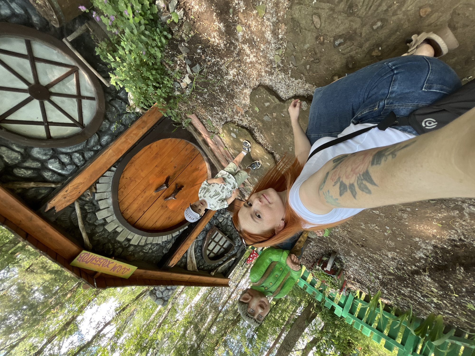 A woman with long red hair and tattoos on her arm is taking a selfie with a young boy and a gnome statue. The boy, wearing a baseball cap and a t-shirt, stands to the right of a wooden door in a fairytale-style house. The gnome, dressed in a green jacket, is positioned to the left of the door. The house has a circular wooden door and a window designed to look like a wagon wheel. A sign above the door reads "ДОМ ХОББИТОВ" (Hobbit House). The scene appears to be in a wooded area, likely part of a park or attraction. The woman is wearing a white t-shirt and blue jeans, and the boy is wearing shorts and sandals. The ground is covered with dirt and small stones, and there is green foliage surrounding the house. This scene is likely located within the city of Chaikovsky, Russia, as indicated by the presence of the "Hobbit House," suggesting a theme park or a specially decorated area.
