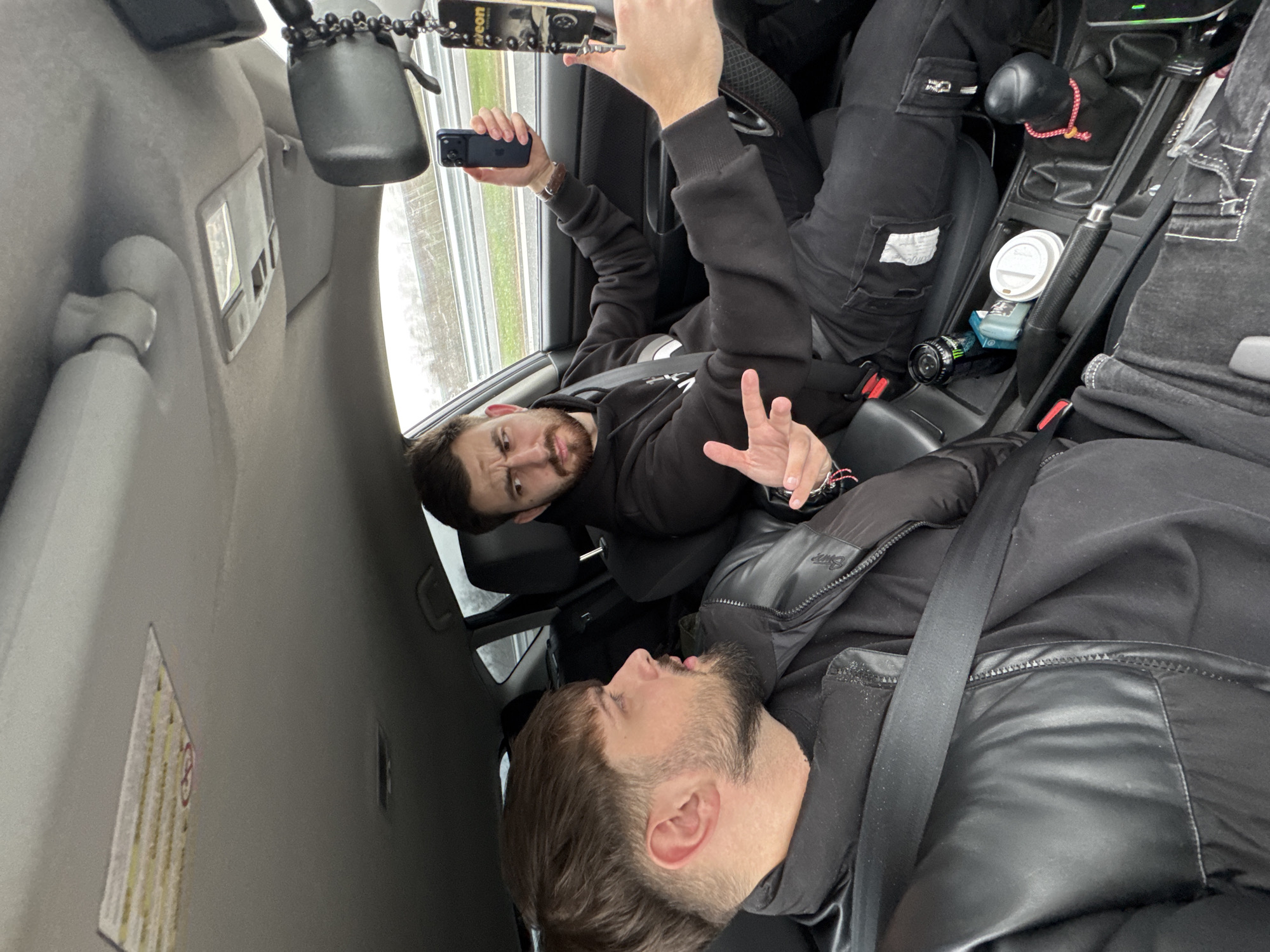 Inside a moving vehicle in Gabarnitsa, Bulgaria, two young adult males are visible. The scene is captured from the rear passenger side, facing forward.
In the front passenger seat, a male with dark hair, a short beard, and a black puffer jacket with a visible seatbelt is turned towards the driver. His mouth is slightly open, suggesting speech or reaction.
The driver, positioned in the left foreground of the frame, is a male with dark hair and a full beard, wearing a black hooded sweatshirt and dark pants. He holds a dark blue smartphone in his right hand, pointed forward and slightly to the right, while his left hand is raised in a gestural pose towards the passenger. His expression indicates engagement, possibly a strong point or reaction. A black rosary-style chain with a crucifix and an "areon" branded air freshener hangs from the rearview mirror.
The car's interior features a dark gray dashboard and upholstery, a light gray headliner, and a center console containing a black Monster energy drink can, a light blue cigarette pack, and a white disposable coffee cup. A gear shift with a black knob wrapped in a red braided cord is also present. Through the front and side windows, a multi-lane road with a guardrail structure and an overcast sky are visible.