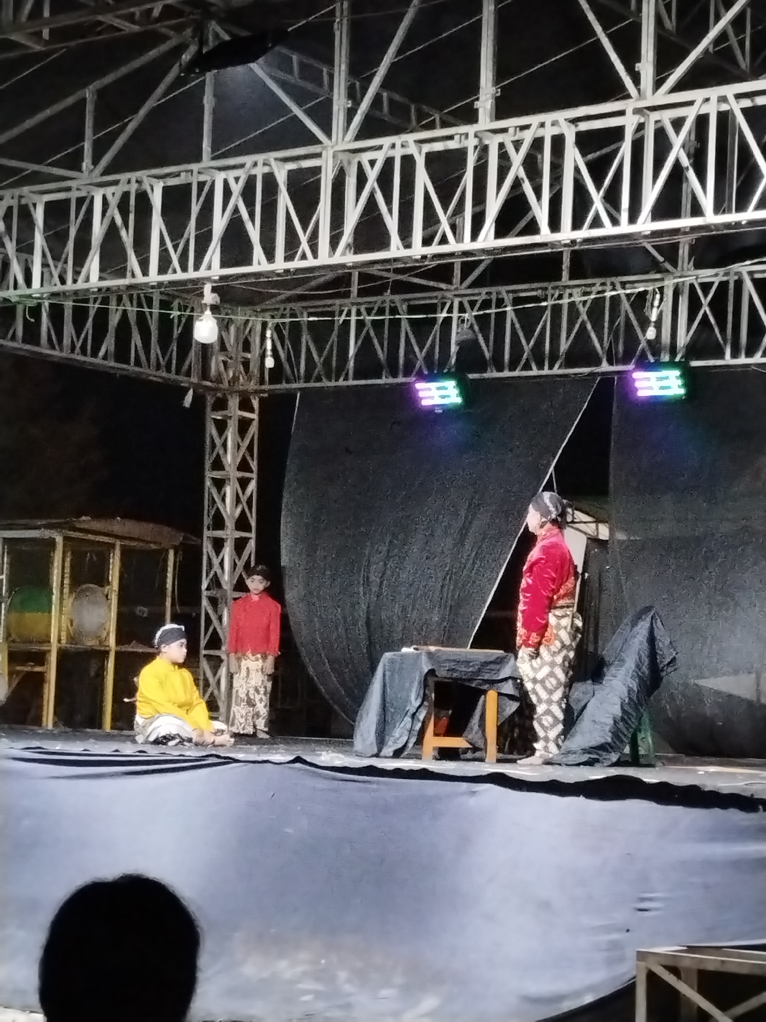 A stage performance is taking place outdoors at night, likely in Kecamatan Pati, Indonesia, given the context. Three individuals are on stage, two of whom are actors in traditional Indonesian attire. One actor, seated on the stage, wears a yellow top and white pants, with a black and white head covering. Another actor, standing, is dressed in a red top and patterned pants, also with a head covering. A third person, likely an adult, stands behind them, their face obscured. The stage is set with a dark backdrop, a table covered with a dark cloth, and two colorful lights illuminating the scene from above. A structure resembling a stage rigging is visible overhead, along with some theatrical equipment and lighting fixtures. In the foreground, the back of an audience member's head is visible, indicating the presence of spectators.