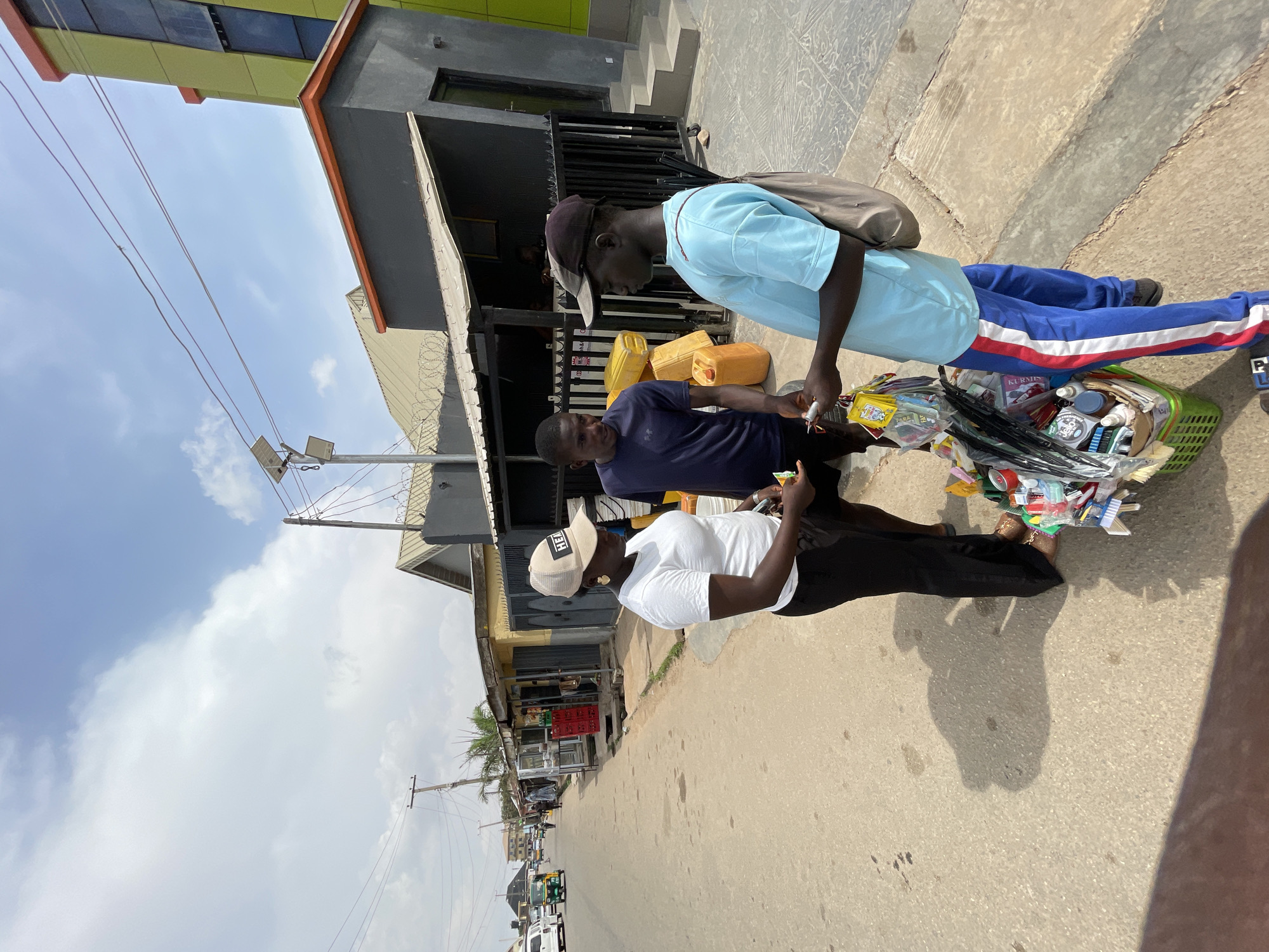 Three men are interacting on a street in Jos, Nigeria. One man, wearing a white t-shirt and black pants, is on the right of the image. A second man is in the middle, wearing a navy blue t-shirt and black pants. The third man, wearing a light blue shirt and blue pants with a white and red stripe, is standing on the left. The man in the light blue shirt carries a basket filled with assorted goods. The basket contains items such as cleaning supplies, food products, and toiletries. The men appear to be engaged in a transaction. Behind them, there is a row of shops and buildings along the street. There are also vehicles and a tricycle (Keke Napep) visible in the background, indicating a bustling commercial environment. Overhead power lines are also present. The sky is partly cloudy. The lighting indicates it is daytime.