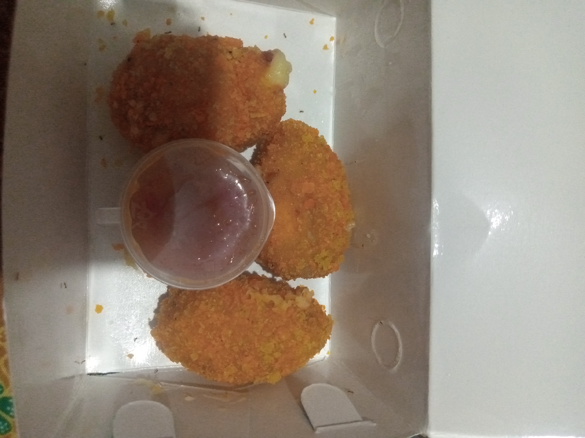 A top-down, slightly angled, full shot shows four fried, breaded food items, likely chicken nuggets or cheese balls, with a small plastic container of red dipping sauce in a white cardboard box. The food items are golden brown and appear to be coated in breadcrumbs, with one item showing a bit of melted cheese oozing out. The dipping sauce is a deep red color and is partially visible in a clear plastic container with a dome lid. The white box has some light brown speckling and a few small black specks scattered throughout. This image was taken in Cianjur, Indonesia.