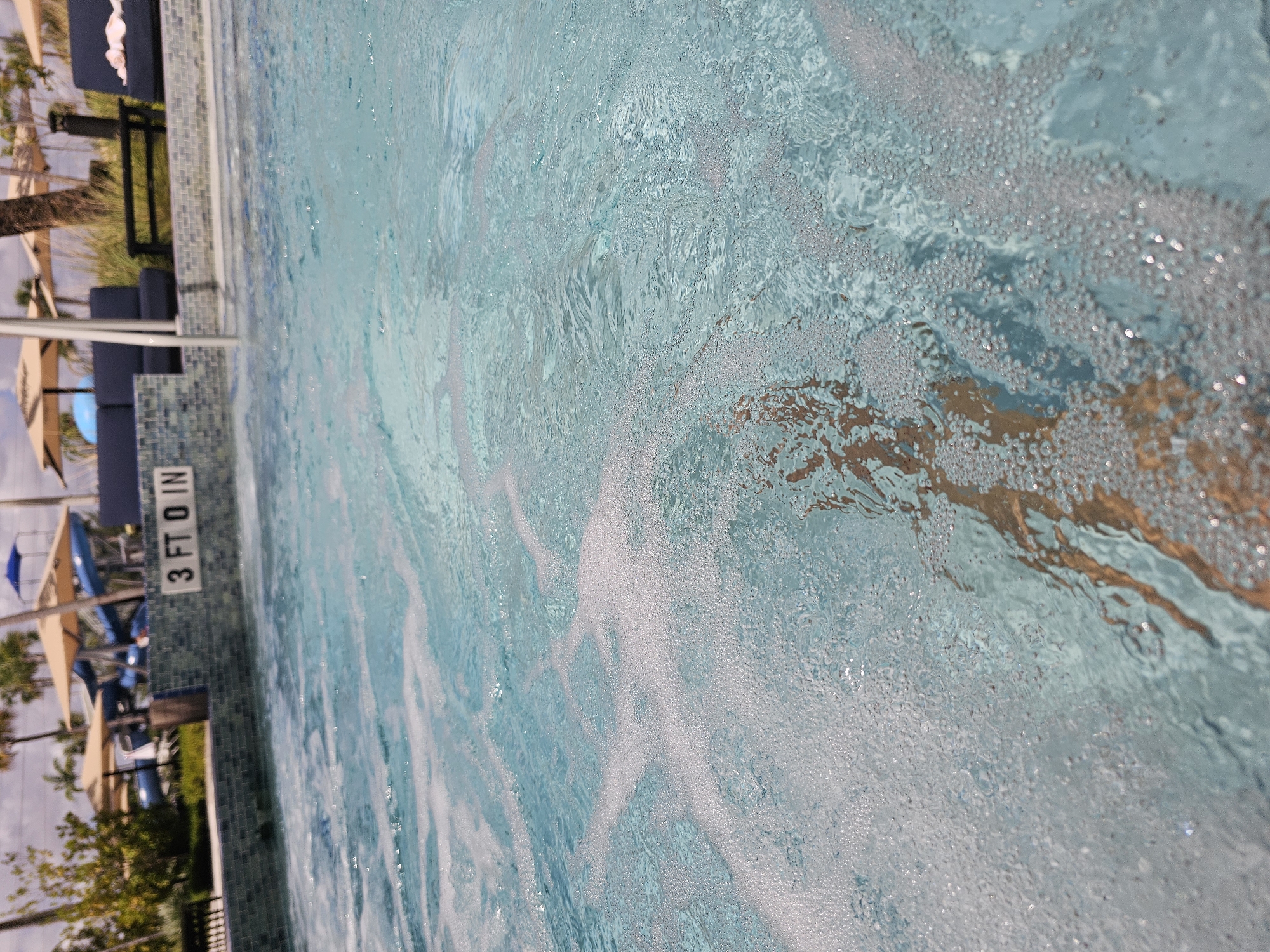 The image depicts a swimming pool with a depth marker of "3 FT 0 IN" on the pool wall. Bubbles and water movement are visible on the surface. In the background, there are lounge chairs with umbrellas, indicating a resort or recreational setting. Based on the style of the scene, the location may be in Miami, United States of America, but this cannot be confirmed with certainty.