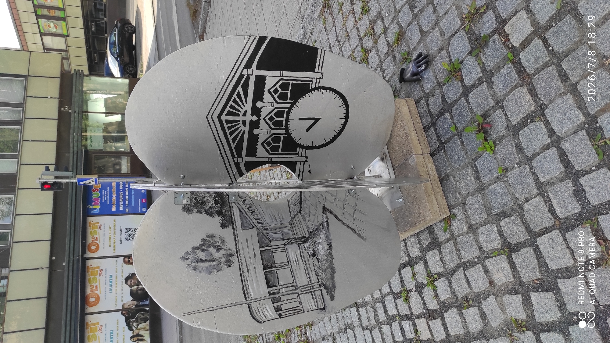 The image shows an outdoor public art installation on a cobblestone street in Iisalmi, Finland. The artwork consists of two large, silver, oval-shaped panels joined by a metal support. Each panel features a black and white drawing. The left panel depicts a building with a covered porch and trees. The right panel shows an architectural facade with arched windows and a large clock face with one hand pointing to approximately 7 o'clock. A black glove lies on the cobblestones to the right of the artwork. In the background, a traffic light is visible, as are posters for events, including "Oluset" and "LIMUSIN." A dark-colored car is parked on the street in the background. The timestamp in the lower right corner indicates July 8, 2026, at 18:29.