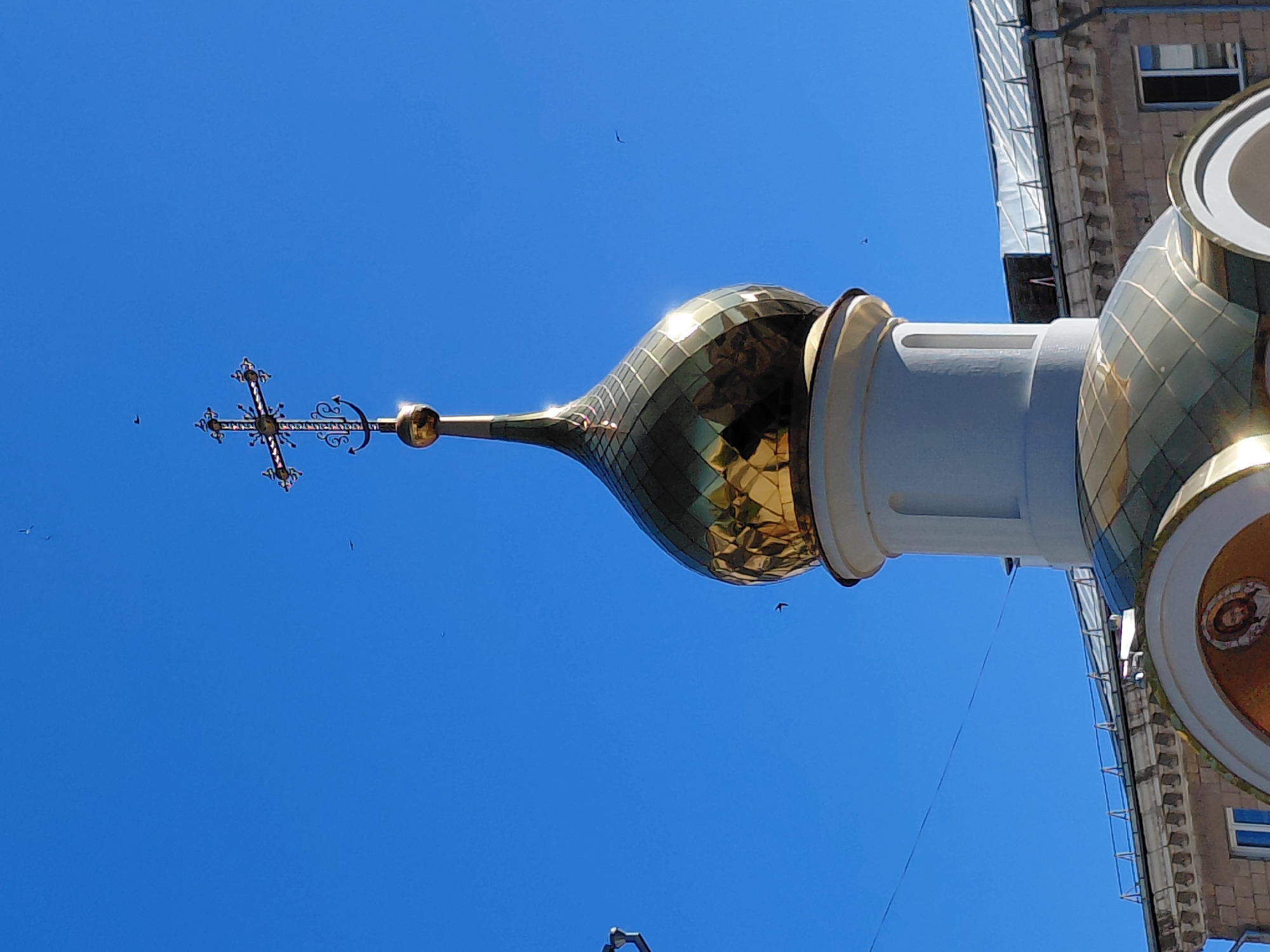 The image shows the top of an Orthodox church dome against a bright blue sky. The dome is gold and faceted, topped with a cross. Several small dark birds are visible in the sky. Below the dome, a portion of a building with a grey stone facade and windows is visible. The location is listed as Unknown, Unknown.