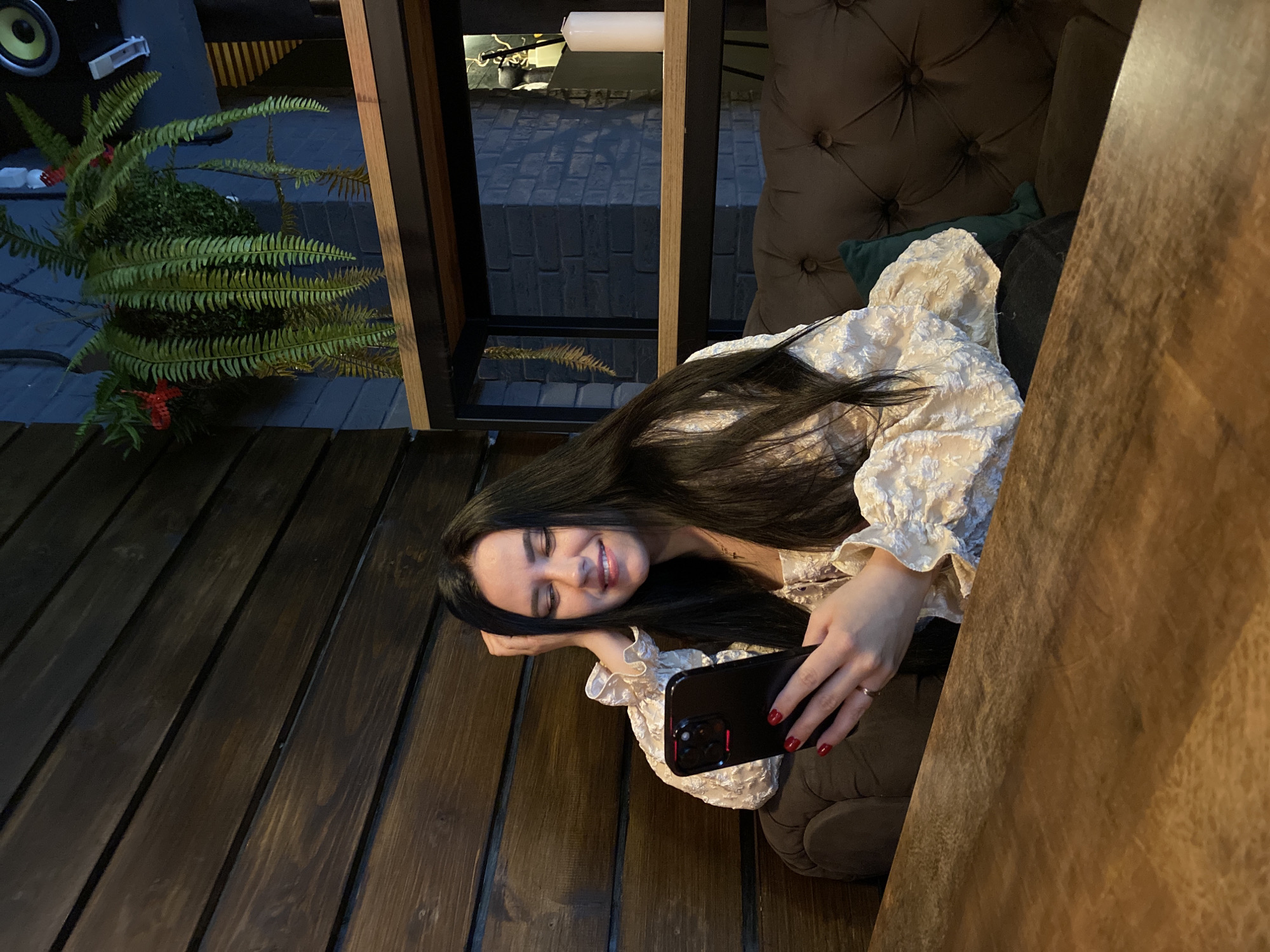 The image, taken in Chaikovsky, Russia, shows a young woman sitting comfortably in what appears to be an indoor cafe or lounge setting. She is smiling broadly with her eyes slightly closed, resting her head on her left hand. Her long, dark hair falls over her left shoulder and arm. She is wearing a cream-colored, textured blouse with ruffled sleeves and dark pants. In her right hand, she holds a black smartphone, likely an iPhone, with her nails painted red.
The setting features dark wooden paneling on the left and extending into the foreground, possibly forming a table or wall. Behind her is a dark, tufted upholstered backrest of a chair or sofa. To the upper left, a vibrant green fern plant with small red accents adds a decorative touch, positioned near a speaker. Through a window-like opening with dark and wooden frames, a view of an outdoor area with a dark, brick-patterned ground is visible, suggesting it is nighttime or late evening due to the absence of daylight. A linear light fixture is also faintly visible in this background section. The overall atmosphere is dim and cozy, with the woman appearing relaxed and happy. No visible text is present in the image.