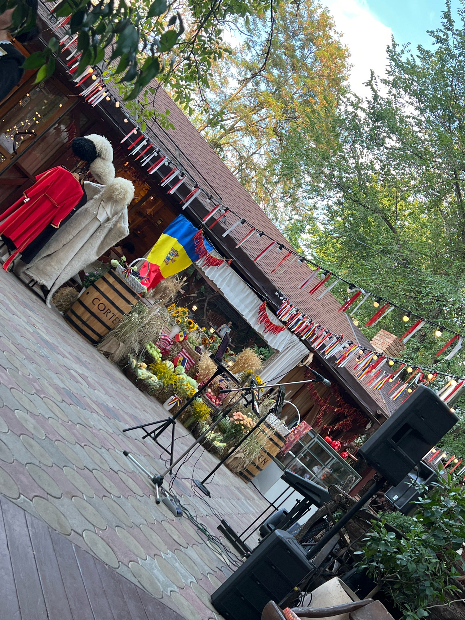 An outdoor scene with a building and trees. The building has a brown roof and is decorated with red and white streamers and small flags. Two traditional outfits, a red robe and a white fur-lined coat with hats, are displayed on mannequins or hangers to the left of the building. In front of the building, a wooden barrel with "CORTE" written on it is placed amongst hay bales, flowers, and produce. A Moldovan flag is visible behind the barrel. The ground is paved with patterned stones. To the right, stage equipment including microphones, speaker stands, and speakers is set up. The overall atmosphere suggests a festive outdoor event, possibly a cultural festival or market.