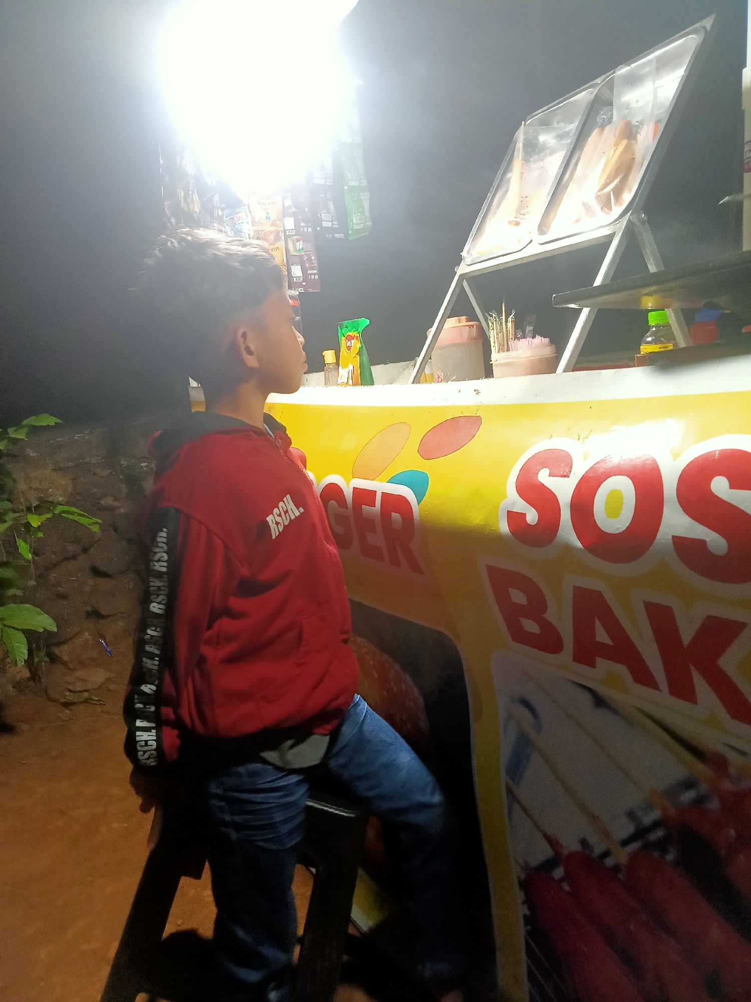 A young boy in a red hoodie stands at an outdoor food stall in Tanggaran, Indonesia. The stall is brightly lit by a large overhead light, casting a strong glow. The boy is looking towards the food displayed on the stall, which includes hot dogs. The stall has yellow signage with red lettering that reads "SOS BAK". The background shows some greenery and a stone wall. The image appears to have been taken at night.