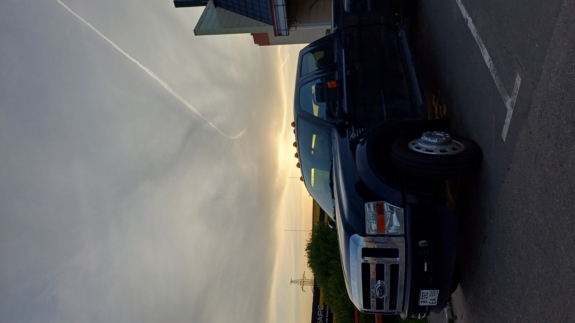 The image shows a dark-colored Ford truck parked on an asphalt surface marked with a white painted parking line. The license plate of the truck reads "B 593 EA 550". Adjacent to the parking spot, there is a building with a pitched roof. Beyond the building and truck, the sky is visible, with a contrail stretching across it. An electrical tower can be seen in the background near some green vegetation. The location is Unknown, Unknown.