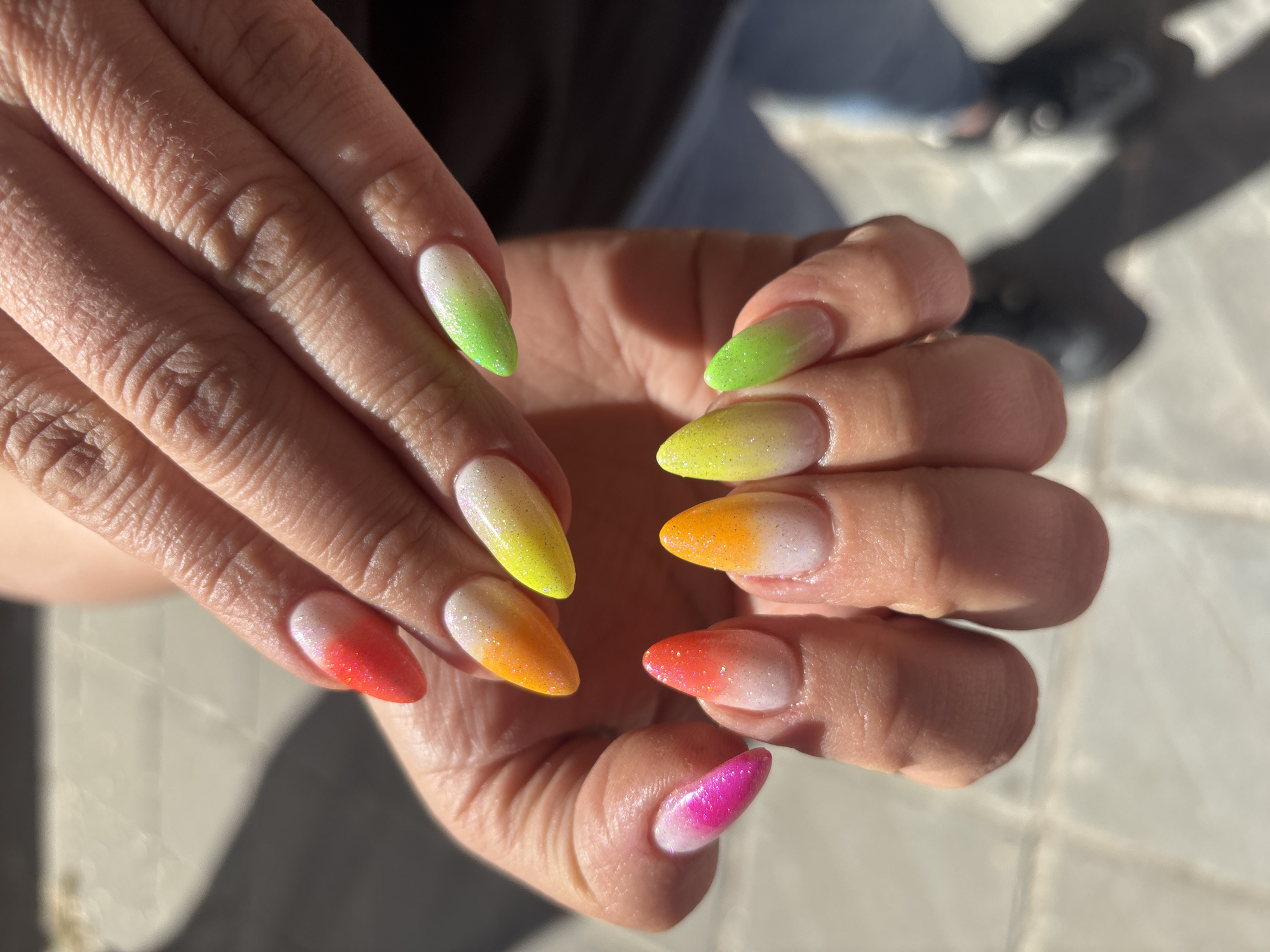 The image captures a close-up of a person's hands, prominently showcasing a vibrant, glittery manicure. The nails are shaped in an almond or oval style, featuring a striking gradient (ombre) design that transitions from a translucent or white base to various bright, sparkling colors at the tips. The colors displayed across the nails form a lively spectrum, including bright green, sunny yellow, warm orange, fiery red, and vivid fuchsia pink. Each nail is coated in a shimmering glitter finish.
The hands are positioned to clearly display the colorful manicure, with fingers slightly spread. The scene is illuminated by strong, direct sunlight, which casts distinct, sharp shadows across the skin of the hands and onto the light-colored, possibly tiled, ground surface below. The background is softly blurred, suggesting an outdoor setting on a clear, sunny day. Visible details on the hands include the natural texture of the skin and some fine hairs. There is no visible text within the image. This scene is observed in Sofia, Bulgaria.