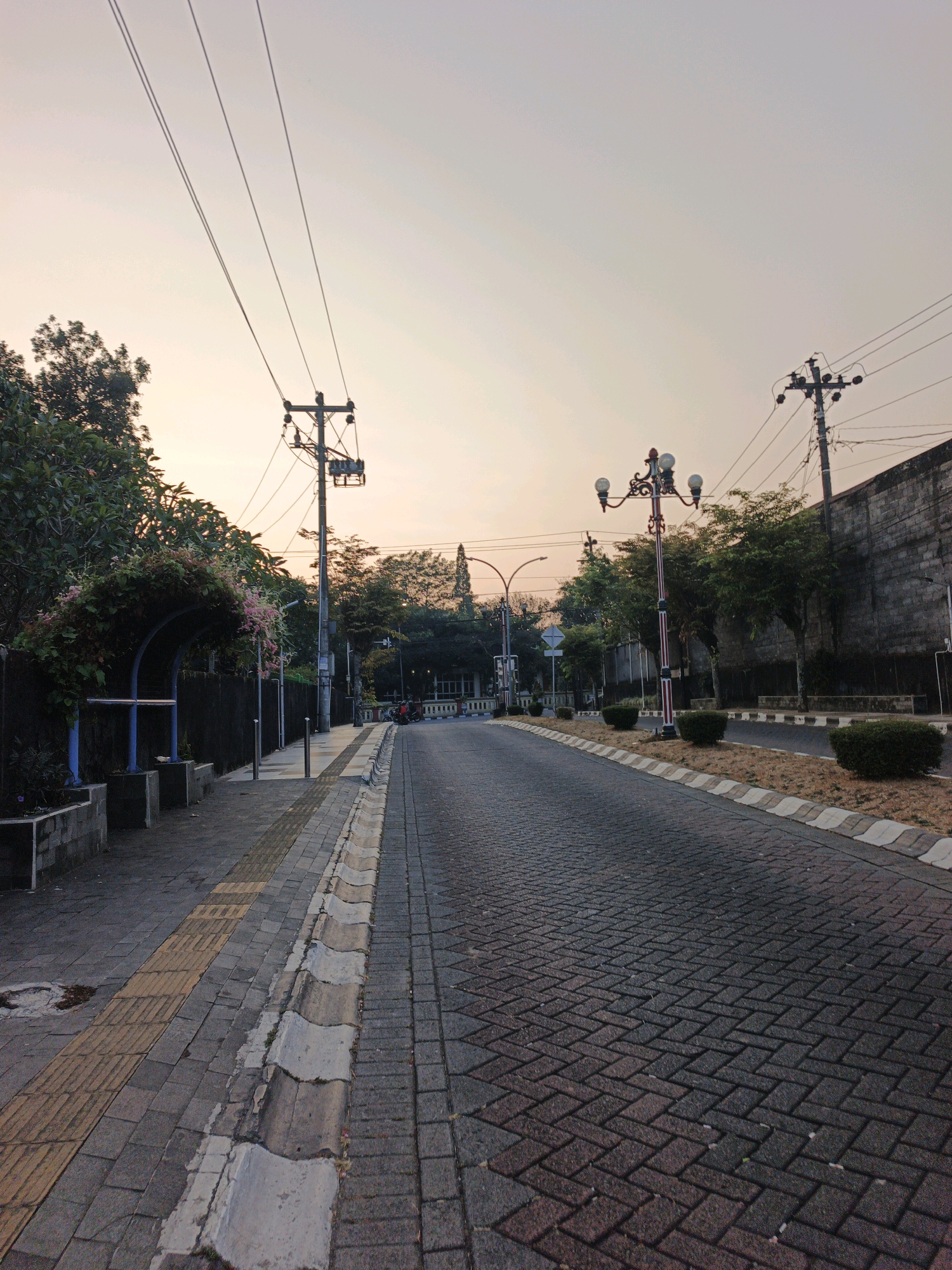 An empty, paved road runs through an urban environment during twilight. The road is constructed of dark brick pavers laid in a herringbone pattern. To the left of the road is a wide sidewalk with textured yellow tiles, suggesting it is a tactile paving area. Beyond the sidewalk is a low wall and lush green foliage, including a decorative archway covered in vines and small pink flowers. Further down the sidewalk, a street lamp with multiple bulbs stands on an ornate, dark pole with red stripes.
On the right side of the road, a narrow strip of dry grass and low, manicured bushes lines the edge. A tall, grey stone wall borders the right side of the scene, with power poles and overhead wires visible against the pale, dusky sky. The sky itself is a soft gradient of pale orange and pink, indicating either sunrise or sunset. No people are visible in the frame, and the scene appears to be devoid of current activity. The overall impression is a quiet, somewhat subdued urban streetscape at the beginning or end of the day.