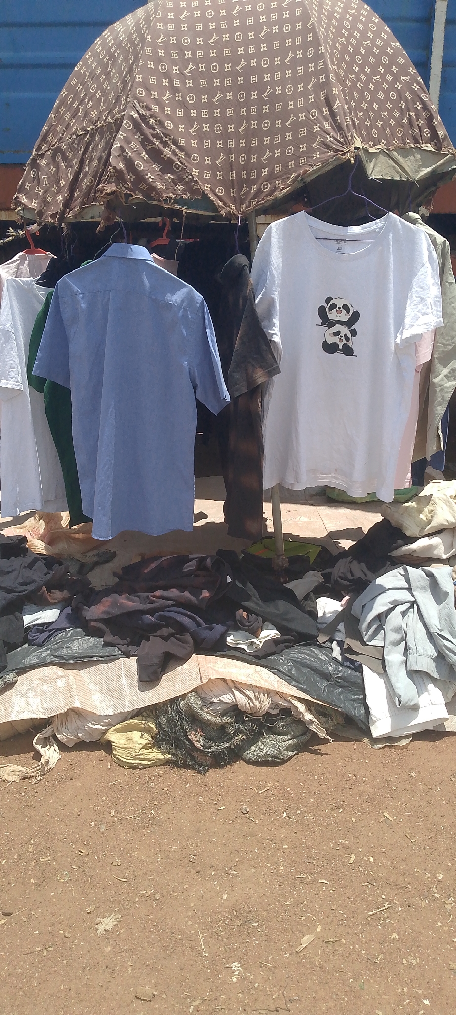 A street market stall is set up outdoors, with various clothing items displayed. A large, brown umbrella with a Louis Vuitton-inspired pattern provides shade. Hanging on display are two t-shirts: one light blue short-sleeved collared shirt, and a white t-shirt with a graphic of two pandas stacked on top of each other. Behind these shirts, more clothing is visible, including white and dark-colored garments. Piled on the ground beneath the hanging clothes are stacks of fabric, including what appears to be blankets or large pieces of cloth. The ground is dirt and debris. The scene depicts a typical open-air market in Tor-Donga, Nigeria, where vendors sell clothing and other goods under makeshift coverings.