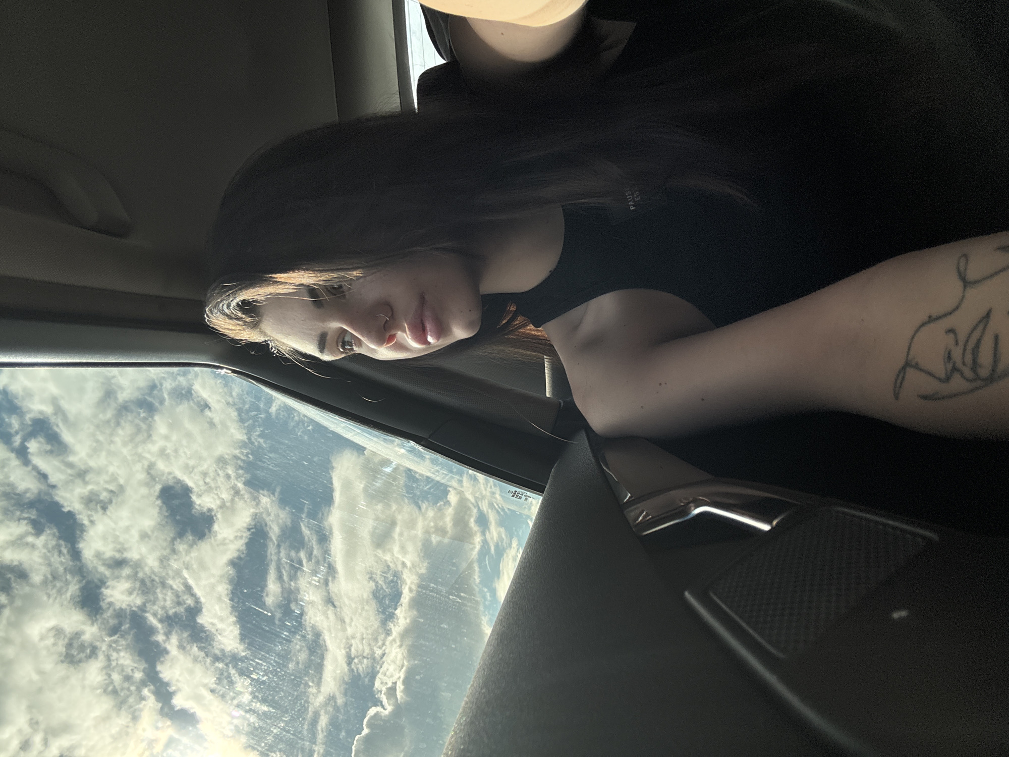 A young person is captured from the waist up, seated inside a car and looking out a side window. The image is oriented vertically, with the person positioned on the left side, her gaze directed towards the bright, cloud-filled sky visible through the window on the right.
The subject has long, dark brown hair, which cascades over her shoulders. She wears a plain black top, and faint text, possibly "PAUSE ES," is visible on the fabric near her right shoulder. Her face is shown in profile, featuring a nose piercing and a neutral, contemplative expression. Visible on her left forearm is a distinct tattoo, rendered in dark lines.
The interior of the car is partially visible, including a dark headliner, a portion of the door panel, and a seatbelt. The sunlight streams in, creating highlights on the person's hair and illuminating the left side of her face.
Outside the car window, an expansive blue sky is filled with numerous large, fluffy white and grey clouds, suggesting a partly cloudy day. The sun is visible towards the bottom of the window frame, casting a bright glow. This scene is set in Herakovo, Bulgaria, during daytime. The person appears to be observing the sky or the external environment.