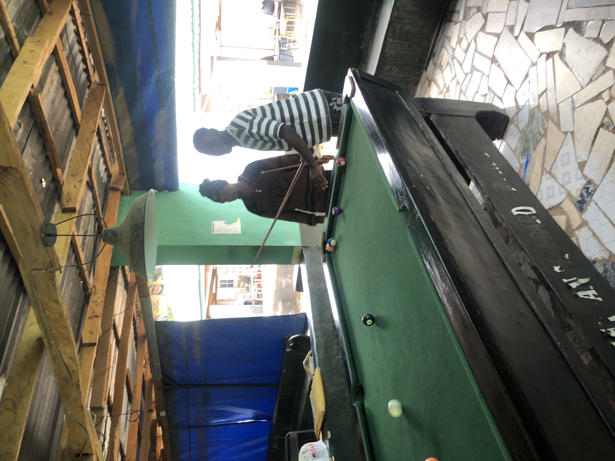 The image depicts two men engaged in a game of billiards within a casually constructed, possibly semi-outdoor or open-air space. The scene is bright, indicating daytime, with natural light entering from an unseen source, supplemented by an overhead, wide-brimmed lamp.
In the foreground, a man wearing a dark, long-sleeved shirt and a dark head covering is leaning over a green-felt pool table, holding a cue stick, appearing to be in the act of taking a shot. His focus is directed towards the pool balls on the table, which include a white cue ball, a solid purple ball, and a red striped ball. To his left, another man stands, observing the game. He is wearing a black and white horizontally striped short-sleeved shirt and faces the viewer.
The pool table has a dark-colored frame, and some indistinct, faded white text is visible on its side, possibly reading "D. A. R." or similar. The floor surrounding the table is covered with an unconventional mosaic of irregularly shaped, broken tiles in shades of white, gray, and blue. Above, the structure features exposed wooden rafters and what appears to be a corrugated metal roof. A large blue tarp is draped over a section of the upper wall or ceiling, possibly providing shade or protection from elements. In the background, beyond the playing area, bright light suggests an open view to the outside, revealing hints of distant structures. A small, crumpled white plastic bag rests on the floor near the table.