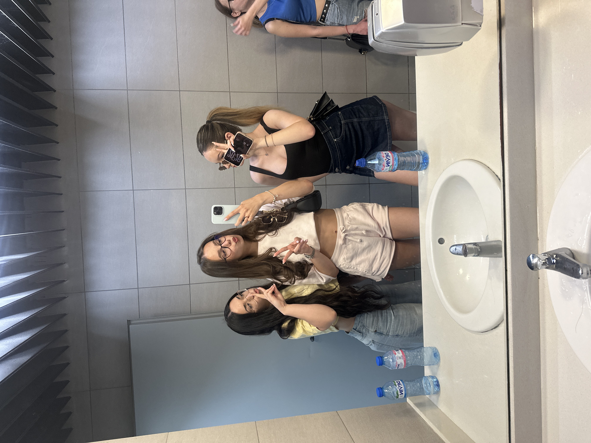 This image depicts a mirror selfie taken by a group of five young women in a public restroom in Blagoevgrad, Bulgaria. The restroom features light grey tiled walls and a large mirror spanning the upper portion of a light-colored countertop. Overhead, the reflected ceiling reveals a distinctive dark grey, slatted or corrugated structure.
In the reflection, from left to right:
A woman with long dark hair, wearing glasses, a yellow short-sleeved crop top, and light blue denim shorts/jeans, is making a "shush" gesture with her right index finger to her lips, looking towards the center of the group.
Next to her, a woman with long brown hair, wearing glasses, a white short-sleeved crop top, and light pink shorts, is holding a white smartphone in her right hand, capturing the mirror selfie. She is making a peace sign with her left hand. Sunglasses are hanging from her top. A black shoulder bag is visible on her left side.
To her right, a woman with brown hair pulled back in a ponytail, wearing sunglasses, a black sleeveless top, and a dark blue denim mini skirt, is holding a dark-colored smartphone in her left hand, which is held up, and making a peace sign with her right hand. A black shoulder bag is visible on her right side.
On the far right, partially visible, is another woman with light brown hair, wearing glasses, a blue sleeveless crop top, and light blue denim shorts. Her right arm is raised, and a black shoulder bag is on her left side.
A fifth woman is partially visible on the extreme left, only her head and upper shoulder reflected.
On the countertop, three clear plastic water bottles with blue caps are visible. Two are in the reflection, and one is on the actual countertop in the foreground. The labels on the bottles are partially visible and reversed. Two white oval sinks with chrome faucets are reflected, and the actual sink with a chrome faucet is visible in the foreground. A silver, rectangular unit, likely a hand dryer or paper towel dispenser, is positioned on the far right of the actual countertop.