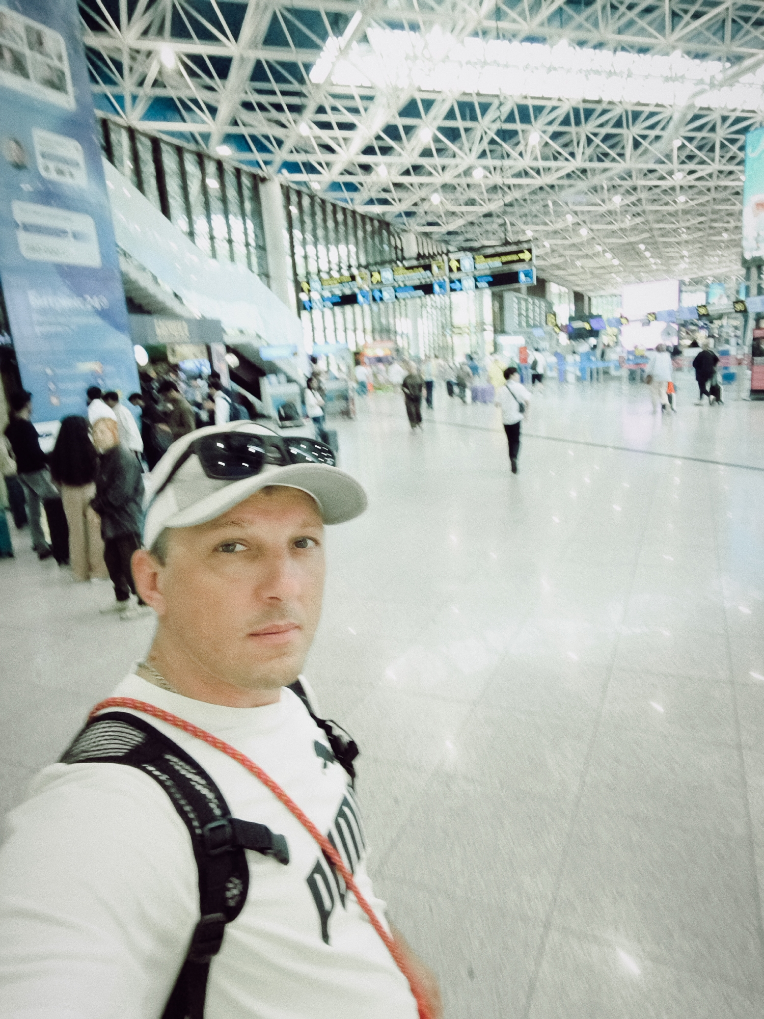 The media file shows a selfie taken inside a modern airport terminal. The main subject is a man wearing a white Puma t-shirt, a baseball cap, and sunglasses perched on the cap. He is also wearing a backpack with a red cord. In the background, other people are visible, appearing to be travelers, some with luggage. The airport features a distinctive geometric ceiling structure with visible lights and trusses, and large windows allowing natural light into the space. Signs with text are present in the background, indicating airport services or directions. The overall scene suggests a busy transit environment. The lighting is bright, typical of an indoor public space, and the floor appears to be polished tile. The image appears to have been taken during the day.