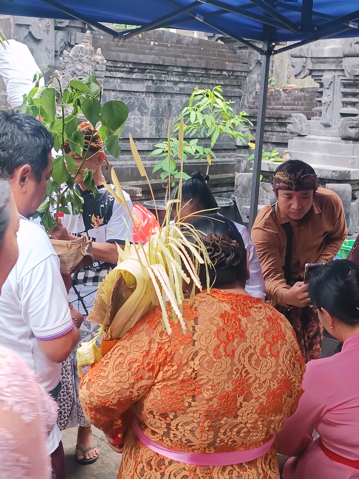 In Tabanan, Indonesia, a group of people are gathered outdoors under a blue canopy. In the foreground, a person wearing an ornate orange lace top with a pink sash stands with their back to the camera, carrying a bundle of palm fronds and other plant materials. To the left, a man in a white t-shirt and a man in a patterned shirt and sarong are visible. To the right, a man in a brown collared shirt and a patterned sarong holds a smartphone, appearing to take a photo. Behind them, stone structures, possibly part of a temple, are visible, with intricate carvings and tiered levels. The overall scene suggests a cultural or religious event.