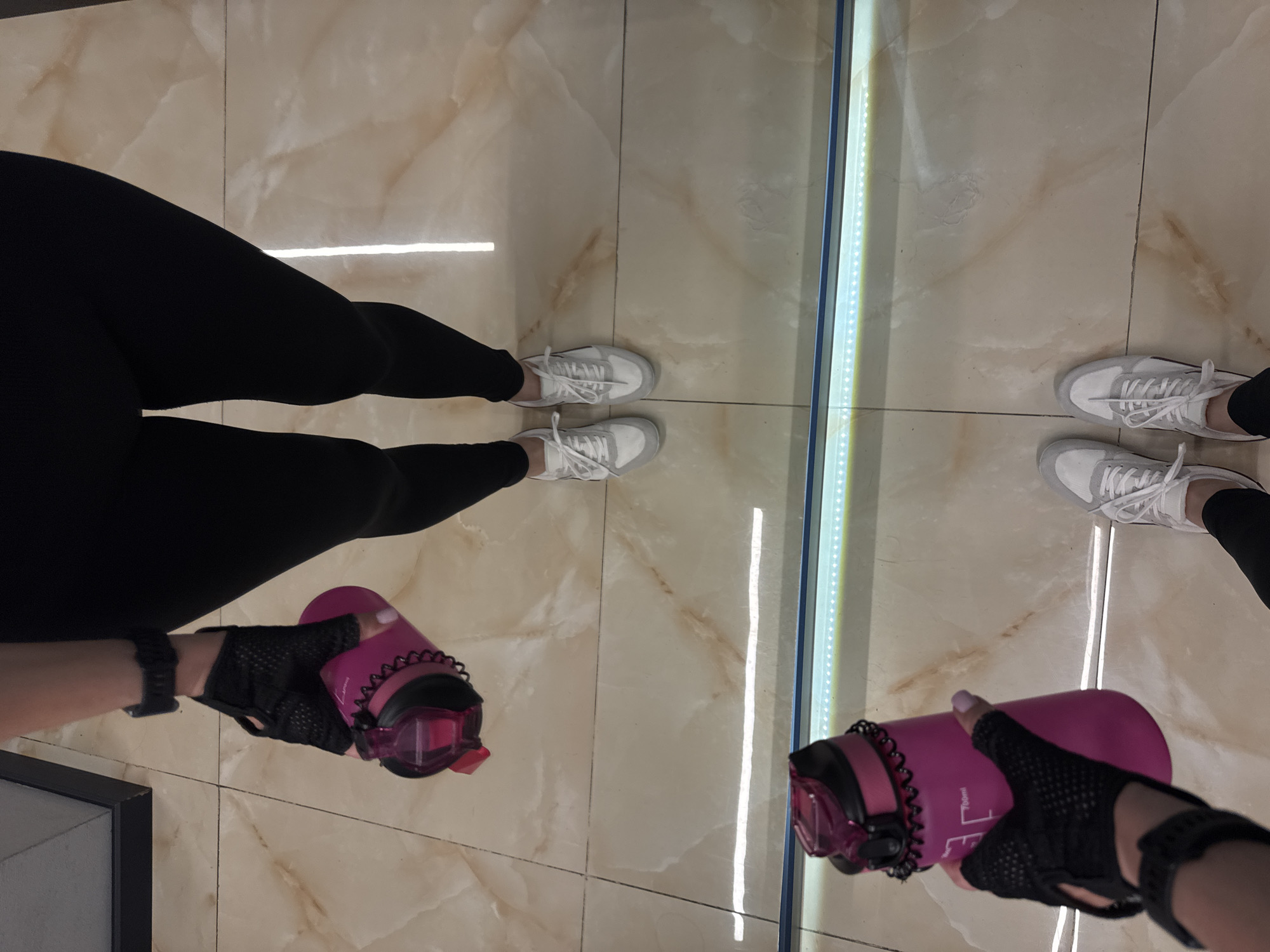 An overhead, slightly angled shot captures a person's lower body, arm, and hand in what appears to be an indoor setting, likely a gym or fitness studio in Sochi, Russia. The individual is wearing black leggings and white athletic sneakers with grey accents. Their left hand, adorned with a black fingerless fitness glove and a black smartwatch or fitness tracker, holds a vibrant pink water bottle with a black lid and a black spiral hair tie around its neck. A reflection of the person's legs, feet, and the water bottle is visible in a mirror on the right side of the frame, separated by a vertical light strip. The floor is covered with polished, light-colored tiles, possibly marble or a similar material, displaying a subtle swirled pattern and grout lines. Partially visible text on the water bottle reads "700ml," indicating its capacity. The scene suggests a moment of rest or preparation during a workout.
