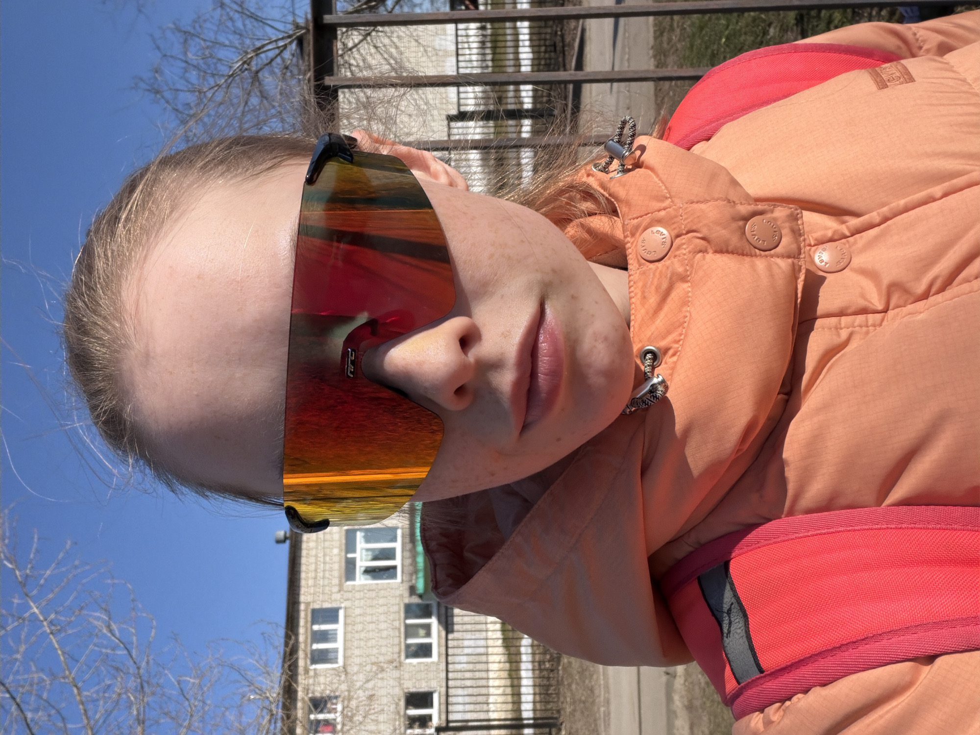 A medium close-up shot depicts a person wearing oversized, orange-mirrored ski goggles and a peach-colored down jacket with red straps from a backpack visible over the shoulders. The jacket has visible buttons near the collar. The person has fair skin with freckles and fair hair. In the reflection of the goggles, there is a cityscape with buildings and bare trees against a clear blue sky. In the background, out of the reflection, there is a light-colored building with multiple windows and a dark fence or railing. Based on the architectural style and the general impression of the buildings, the image is likely from Berezniki, Russia. The photograph is taken during daylight hours on a clear, sunny day.