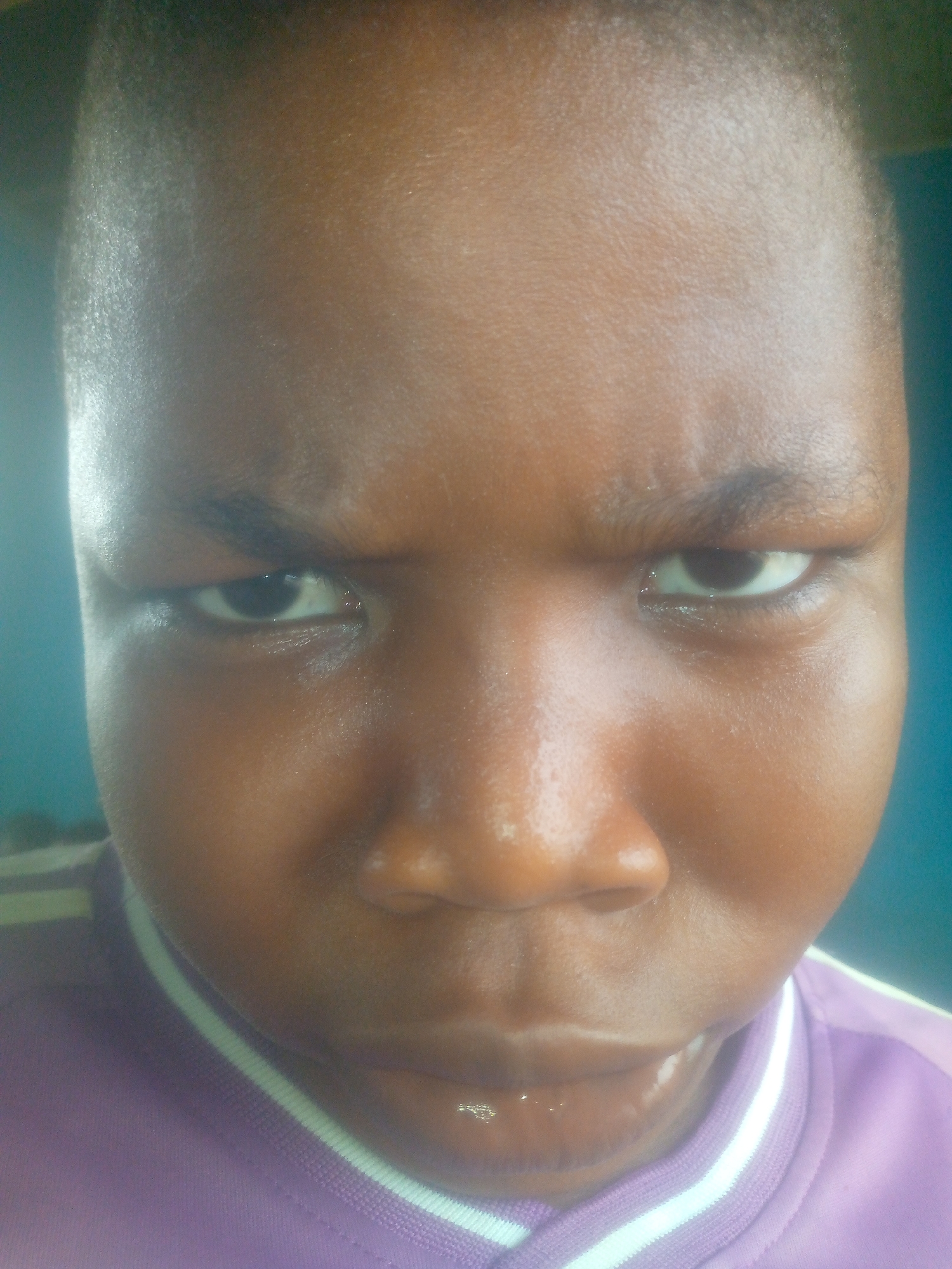 A close-up photograph depicts a person with dark skin and short hair. The person's facial expression appears stern, with furrowed brows and a focused gaze. The individual is wearing a purple shirt with a white-striped collar. The background is indistinct, with a blueish hue. The location is unknown.