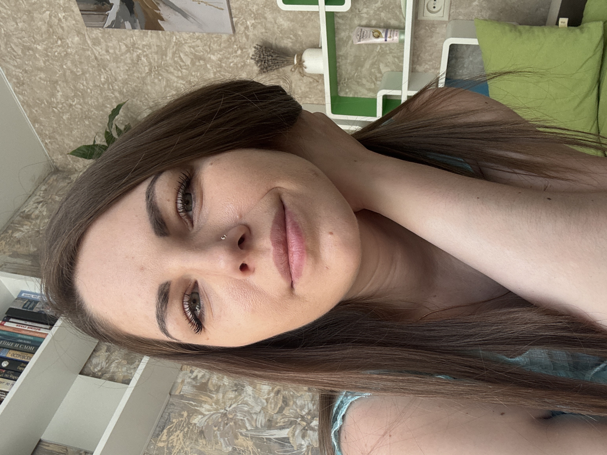 A close-up shot depicts a woman with long brown hair, fair skin, and a nose piercing. She is holding her hair with one hand. Her lips are full and her eyes are open, looking at the camera. The background includes a patterned wall, a white bookshelf filled with books, a decorative white shelf with a green accent, and a green pillow. The location is unknown, although it might be Раздолье, Russia.