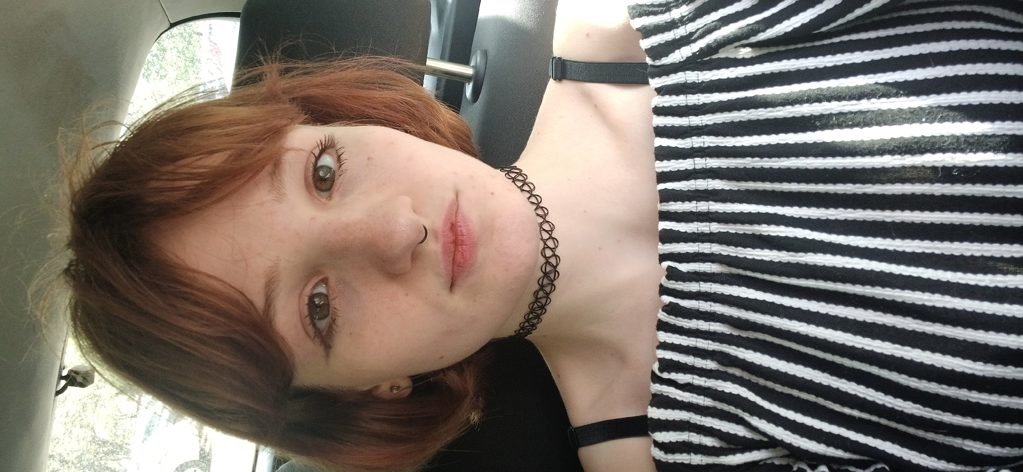 A young, pale-skinned woman with light brown hair is in the driver's seat of a vehicle, likely a car. She is wearing a black and white striped shirt and a black choker necklace, plus a black band around her wrist. She has freckles on her face and a ring in her nose. The environment is the interior of a vehicle, possibly a car. Outside of the car window is light foliage, though the location in the city of Bar, Montenegro cannot be determined from the image.