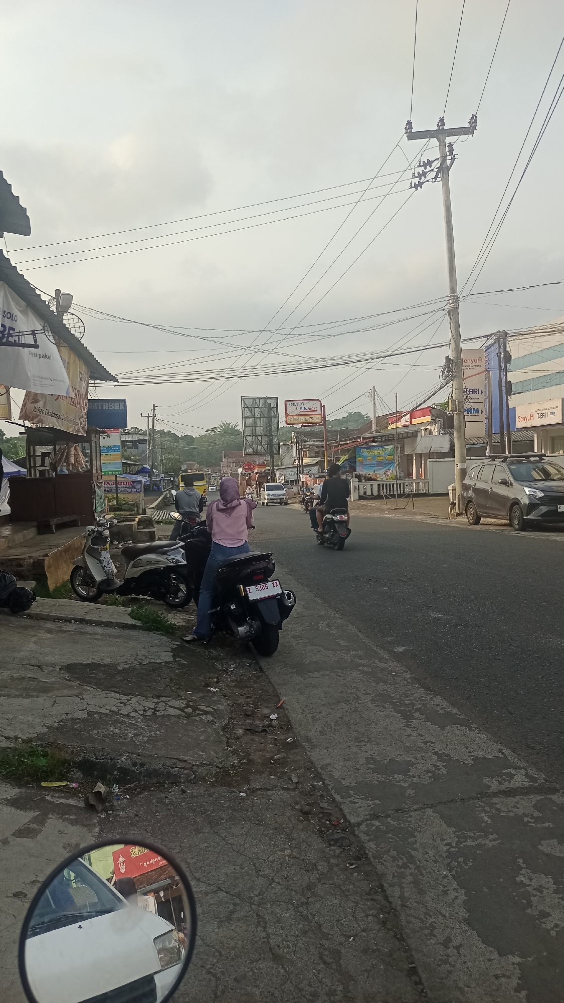 The image depicts a street scene in Karyamandala, Indonesia, during daylight hours with an overcast sky. In the foreground, a gray concrete sidewalk runs along the left side of a paved road. Several motorcycles are parked on the sidewalk. One motorcycle, a dark color with a license plate reading "Z 5385 XX," has a person wearing a pink hooded jacket and blue jeans sitting on it, facing away from the camera. Another lighter-colored motorcycle is parked to its left.
On the road, several vehicles are present. Two people are riding a black motorcycle, moving away from the camera. Further down the road, a gray car is visible on the right side. The road is lined with buildings and shops on both sides, displaying various signs and advertisements, including one that appears to be for BRI. Utility poles with numerous wires crisscross the sky. In the reflection of a motorcycle's rearview mirror in the bottom left corner, a white vehicle and part of a building with red lettering can be seen. The environment appears to be a commercial or semi-urban area.