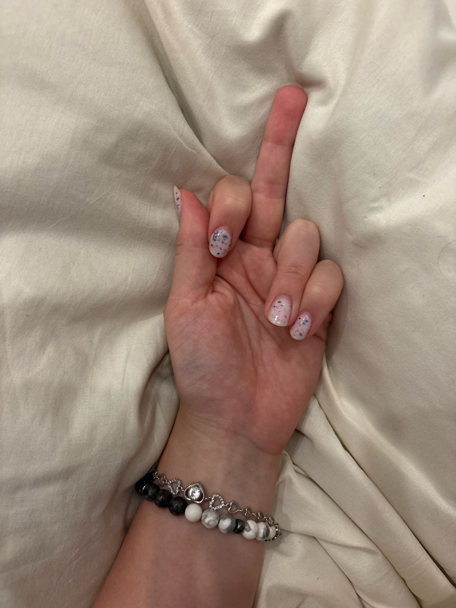 A person's hand with painted nails is raised with the middle finger extended, resting on a wrinkled cream-colored fabric. Two beaded bracelets are worn on the wrist. The image is an indoor shot.