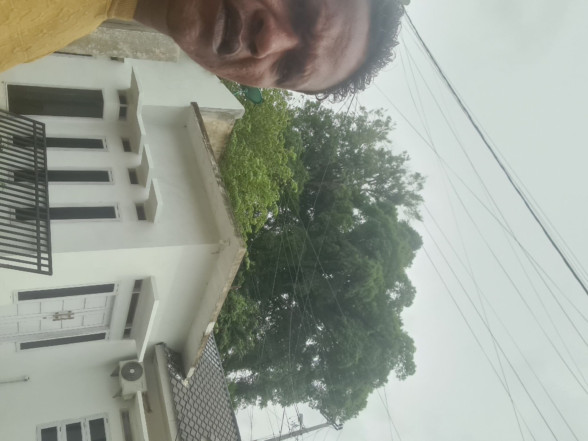 The image shows a close-up selfie of a man with dark skin and curly hair, wearing a yellow sweater. He is looking directly at the camera with a neutral expression. Behind him, a white house with a dark-tiled roof is visible. The house has several windows, some with black frames, and a white double door. An air conditioning unit is mounted on the exterior wall. Power lines crisscross the overcast sky above. In the background, lush green trees, including a very large one, dominate the upper portion of the image, suggesting a residential area possibly bordering a natural landscape. The scene appears to be in Lhokseumawe, Indonesia, indicated by the context provided.