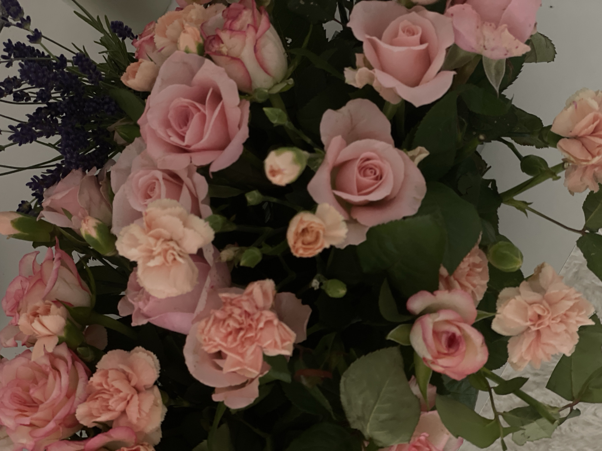 The image is a close-up of a floral arrangement featuring various shades of pink roses and carnations, interspersed with sprigs of dark purple lavender. The flowers are densely packed, with green foliage visible throughout. The lighting is soft, casting gentle shadows and highlighting the delicate textures of the petals. The composition is focused on the blooms, with a blurred background that suggests an indoor setting. There is no visible location information within the image to determine its presence in the city of Ulmen, Germany.