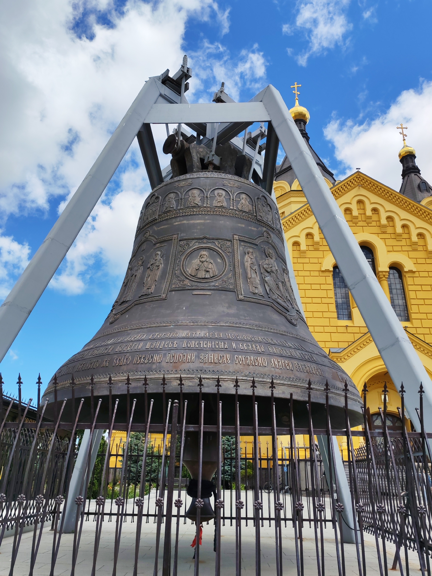 A low-angle, outdoor shot captures a colossal bronze bell on a metal A-frame, dwarfed by a vibrant yellow church with golden domes under a bright blue, partly cloudy sky. The bell, heavily decorated with bas-reliefs of figures and intricate patterns, is encircled by a dark, ornate metal fence. The church's architecture is distinctly Russian Orthodox, featuring arched windows and decorative elements. The scene suggests a sunny day, possibly midday given the brightness of the sky. The bell appears to be a historical or religious artifact, possibly a monument or part of a functioning place of worship. The text inscribed on the bell is visible, though partially obscured, and appears to be in Cyrillic script. The overall impression is one of grandeur and historical significance. The bell's imposing size and the surrounding religious architecture evoke a sense of reverence and awe.