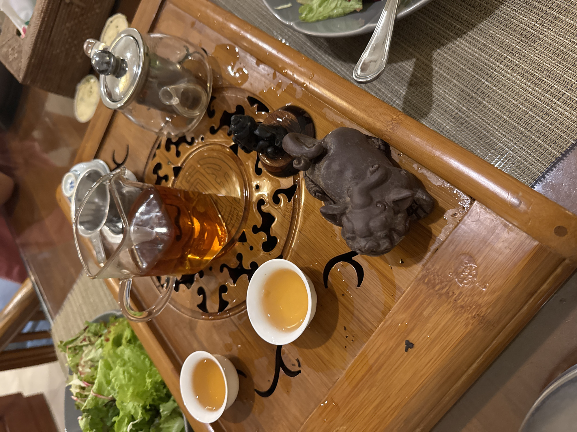 The image shows a tea set on a wooden tray. A glass teapot with brown liquid is tilted, as if pouring tea into two small, white cups. A second glass teapot and a small, sculpted figure of an animal are also on the tray. Spilled water droplets are visible on the tray's surface. The tray has a patterned cut-out design. Part of a salad dish with silverware is in the upper right.
The scene appears to be indoors, on a table with a neutral-colored placemat. There is no specific indication of it being in Kuta Selatan, Indonesia, or any people present.
