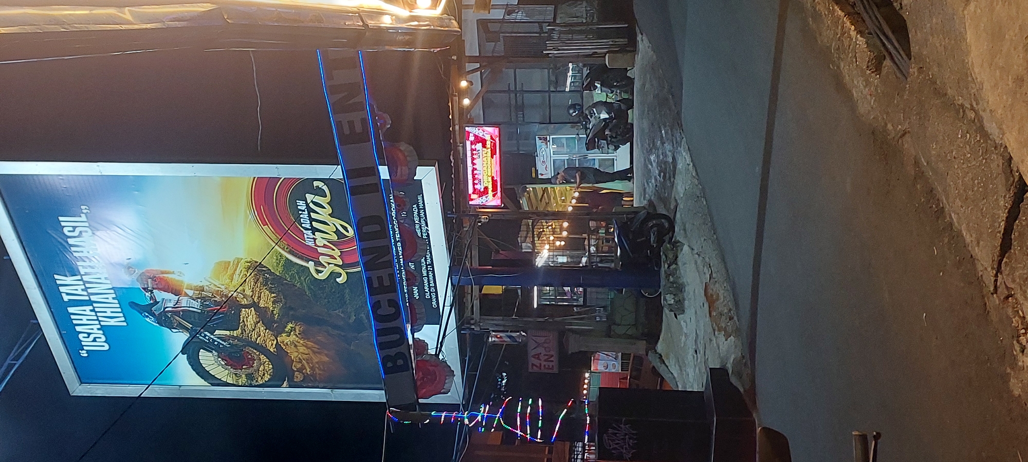 The image depicts a street scene at night in Jayapura, Indonesia. Dominating the upper portion of the frame is a large billboard featuring a motocross rider on a cliff with the text "USAHA TAK KHIANATI HASIL" (Effort Does Not Betray Results) and "KITA ADALAH SURYA" (We Are Surya). Below the billboard, a sign with blue neon lights reads "BUCEND II ENT".
In the middle ground, a dimly lit establishment is visible with several motorcycles parked outside. A person stands near the entrance of this establishment, holding a small object, possibly a phone or a menu. Another person is partially visible inside. The lighting suggests a commercial area, possibly a restaurant or shop.
The foreground shows a street with a textured pavement. To the left, a dark, rectangular object with graffiti is present, and a string of colorful festive lights hangs diagonally. The overall atmosphere is that of a bustling urban environment at night, illuminated by artificial lights and the glow from the billboard and establishment.