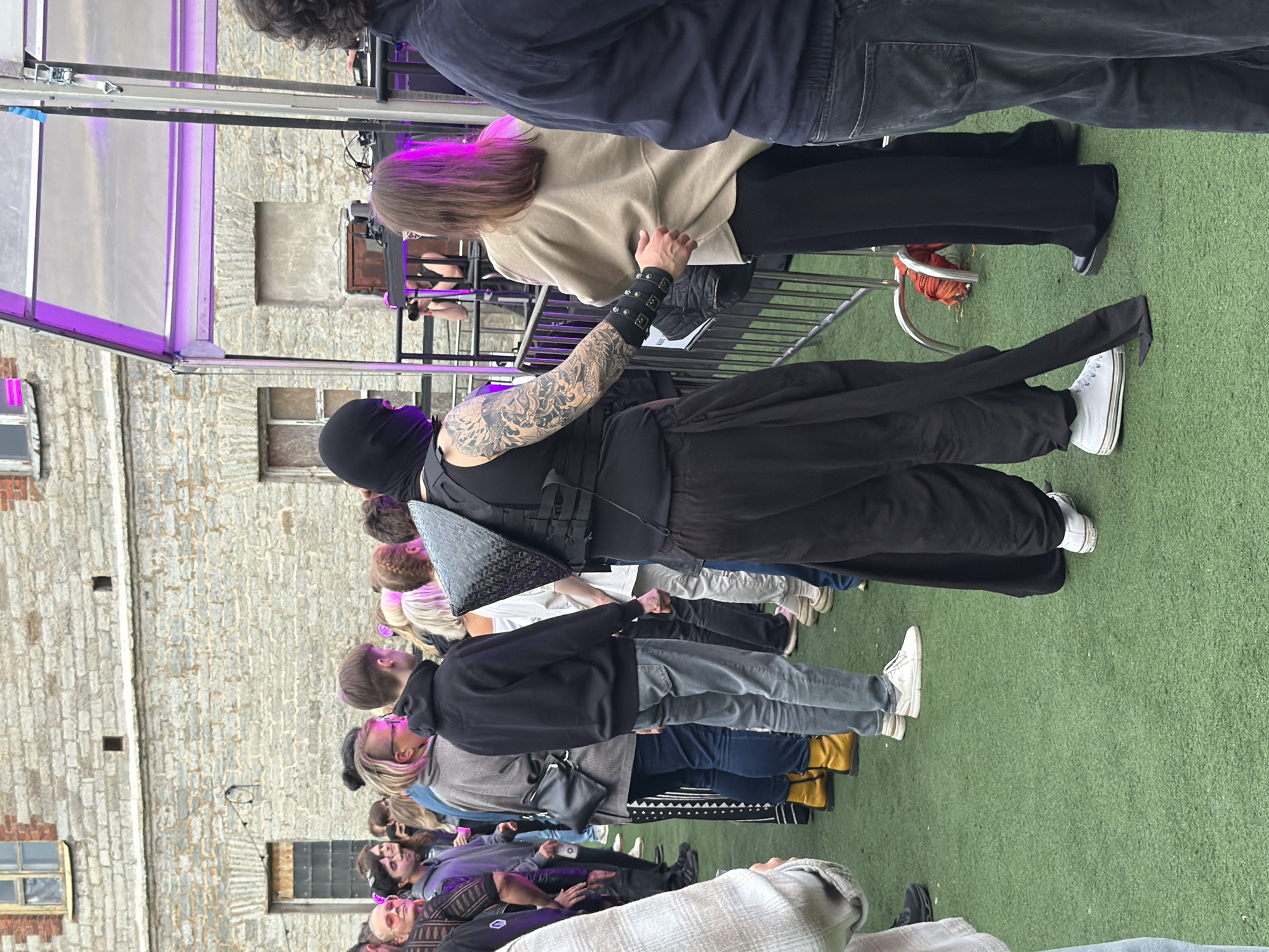 The image shows a crowd of people gathered outdoors on a patch of green turf, likely artificial grass, in an urban setting. In the background, a weathered brick building with visible windows and architectural details suggests an older style of construction. A purple-lit canopy structure with metal supports is present on the right side of the image, possibly indicating a stage or an event setup.
A man in the foreground is prominently featured, wearing a black balaclava that covers his head and face, along with a black vest that has a distinctive metallic, angular shoulder piece. He has extensive tattoos visible on his left arm. His attire includes loose-fitting black pants and white sneakers. He is holding the arm of a person to his right, who is wearing a light-colored sweater and dark pants. This interaction suggests a close proximity or a gesture of guidance or familiarity.
Other individuals in the crowd are seen standing in a line or cluster. Their clothing is varied, with some wearing casual attire like hoodies and jeans, while others are dressed in darker clothing. One person towards the left is wearing bright yellow boots. The crowd appears to be facing a particular direction, possibly towards the purple-lit structure. The overall atmosphere seems to be that of an outdoor gathering, potentially a concert, festival, or public event.
The presence of the brick building and the overall architectural style, combined with the mention of Tallinn, Estonia, points to a location within the historic part of the city, perhaps an old industrial area that has been redeveloped for cultural events.