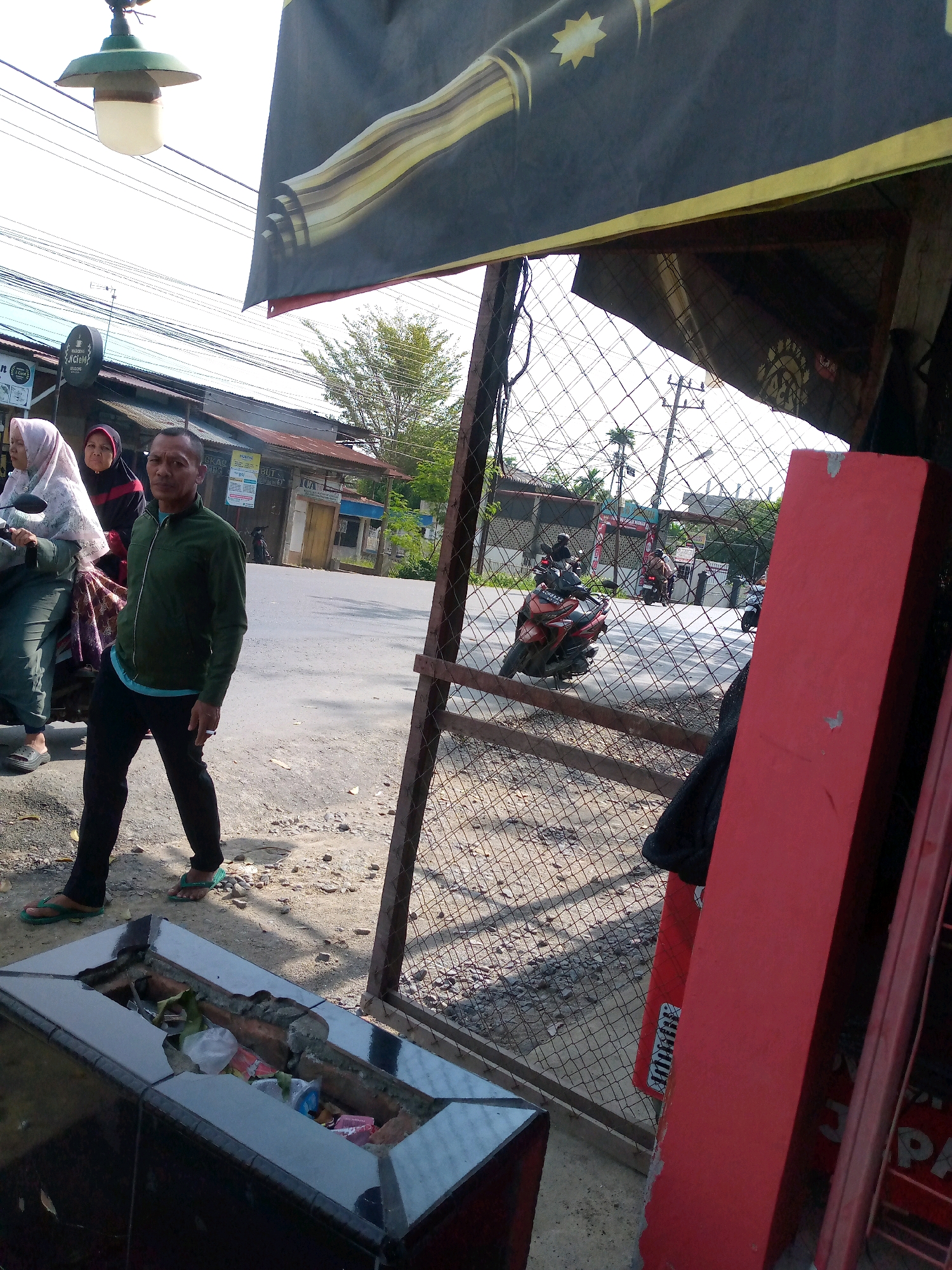 A street scene in Langa, Indonesia, features a man in a green jacket and black pants walking in the foreground, with two women in hijabs on a motorcycle behind him. A large black banner with yellow and gold wavy lines and a starburst graphic hangs from an overhead structure, partially obscuring a green street lamp. The street is paved with asphalt and has a dirt and gravel shoulder. A metal fence with diamond-shaped mesh encloses an area to the right, where a tall, red rectangular object stands. In the background, there are buildings with corrugated metal roofs, trees, and utility poles with wires. Several motorcycles are visible on the street. A dark, rectangular object with a broken top, containing trash bags and plastic, is positioned in the lower left corner of the image. The lighting suggests it is daytime, with sunlight casting shadows.