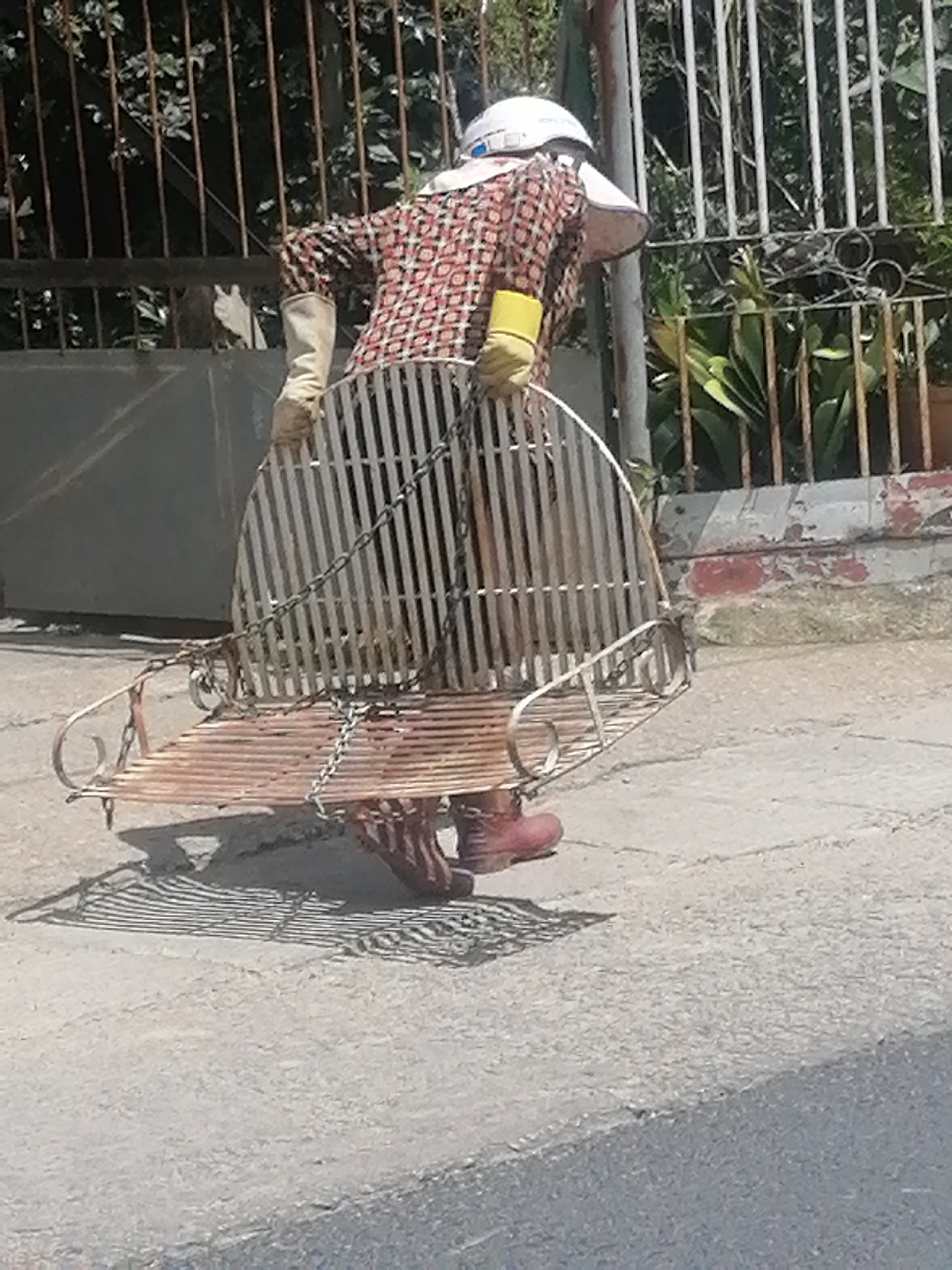 A person wearing a patterned shirt, white hard hat, and yellow gloves is carrying a metal swing set in a street in Da Lat, Vietnam. The swing set is made of metal and has a curved back and seat. The person is walking on a paved surface, and there is a fence in the background. The person appears to be moving the swing set from one location to another.