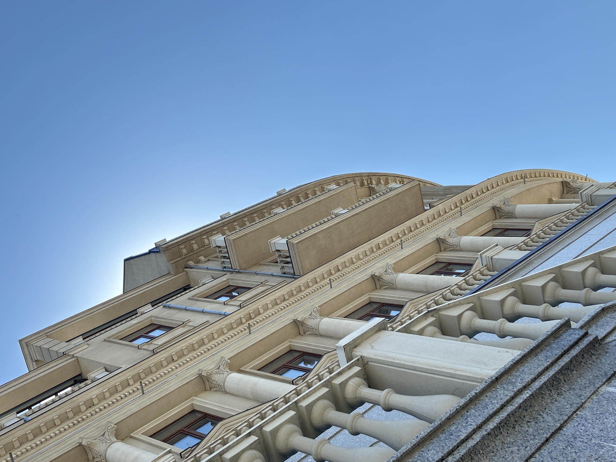 The image shows a low-angle view of a building in Tiraspol, Moldova. The building is made of light-colored stone and features ornate architectural details, including columns, balustrades, and decorative moldings. The windows are rectangular with dark frames. A clear blue sky occupies the majority of the right side of the image. The sun is partially obscured by the upper part of the building, creating a bright glare. The left side of the image is dominated by the building, which appears to be several stories high and has a curved facade. The lower portion of the building visible in the foreground is clad in grey stone with a speckled texture, while the upper sections are a uniform light cream color. The building has balconies with decorative railings.