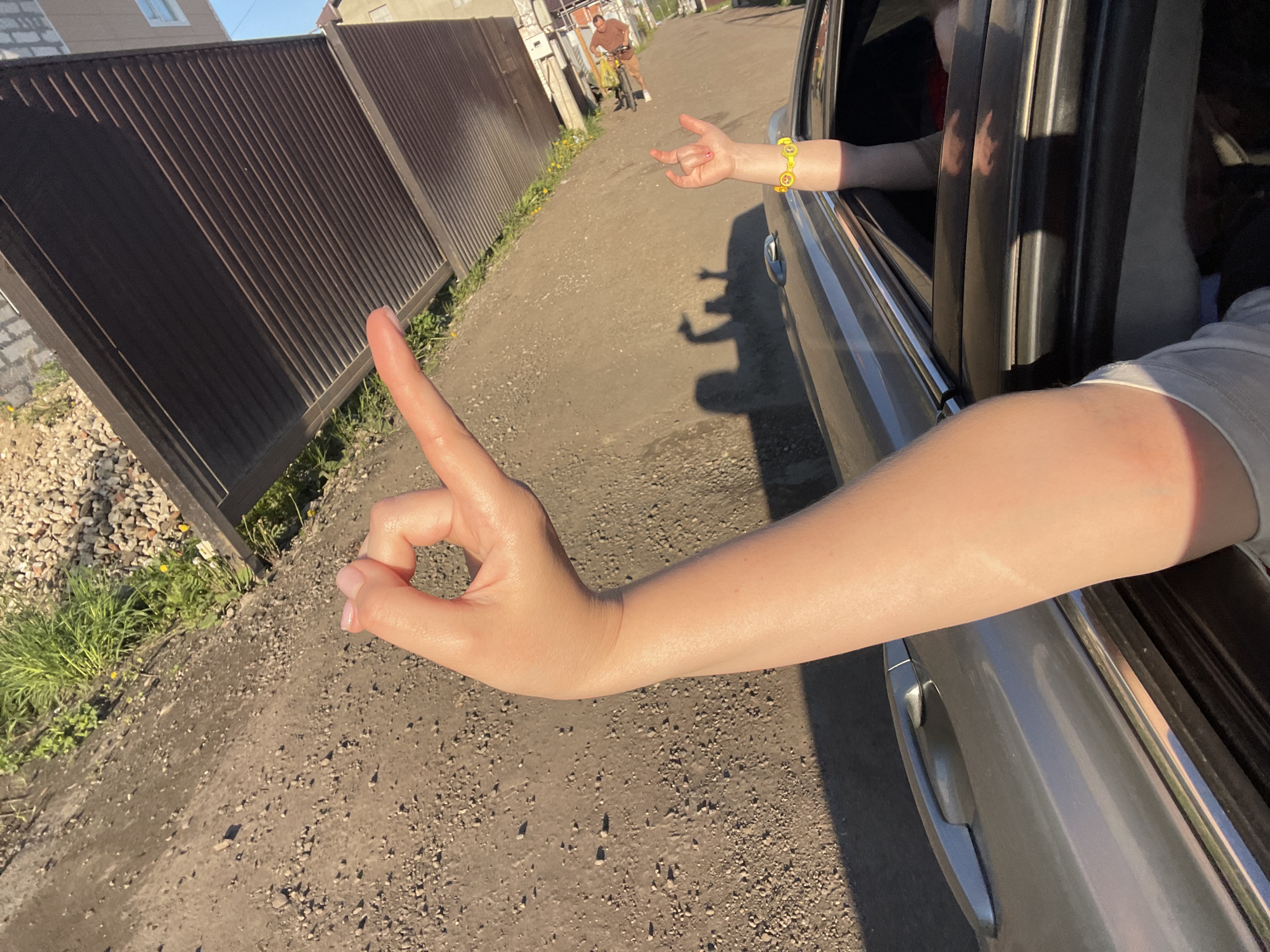 A daytime, eye-level shot shows the arm of a person extending from the window of a silver car. The person is making a hand gesture with a raised index finger and a curled thumb. The arm exits the car window from the lower right, and their hand is in the lower left, appearing to point towards a brown metal fence on the left side of a dirt road. A second arm, also extending from the car window, wears a yellow bracelet with a pattern of smiley faces.
In the background, a person on a yellow bicycle rides away from the camera, down the dirt road. Residential structures are visible along the road, one with a light-colored exterior.
The location is likely Molokovo, Russia, as it aligns with the information given in the prompt.