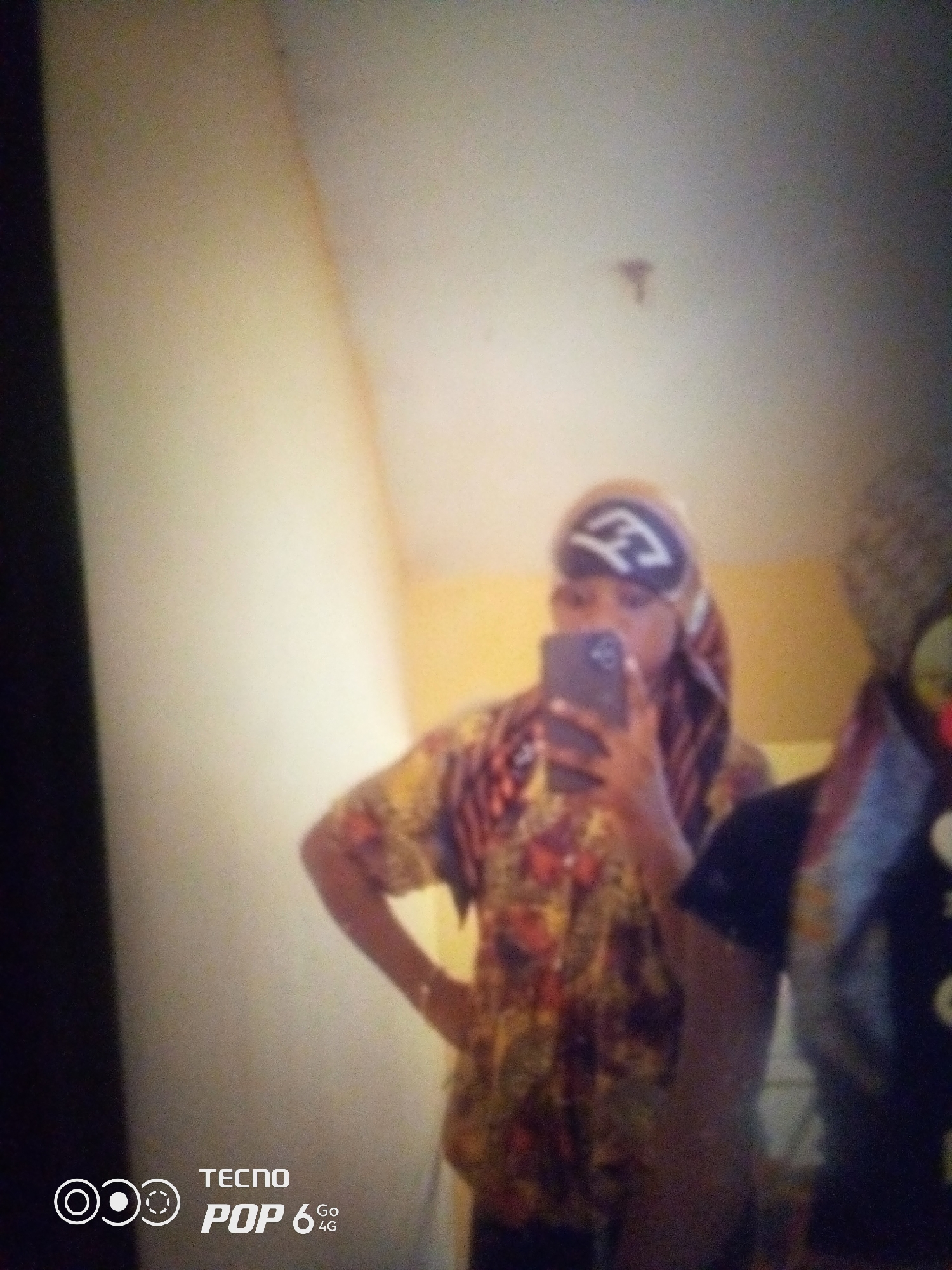 A blurred, waist-up, full-body shot shows a person standing in front of a light-colored wall, taking a selfie in a mirror. The person is wearing a headscarf with a white design and a colorful, patterned shirt. They are holding a dark-colored smartphone in their right hand. Another person is partially visible to the right of the frame. The bottom left corner of the image displays the "TECNO POP 6 4G" logo. The setting appears to be indoors, with a yellow wall in the background. The image quality is low, with significant blur and graininess. The specific location in Howayi, Nigeria, cannot be determined from the visual information provided.