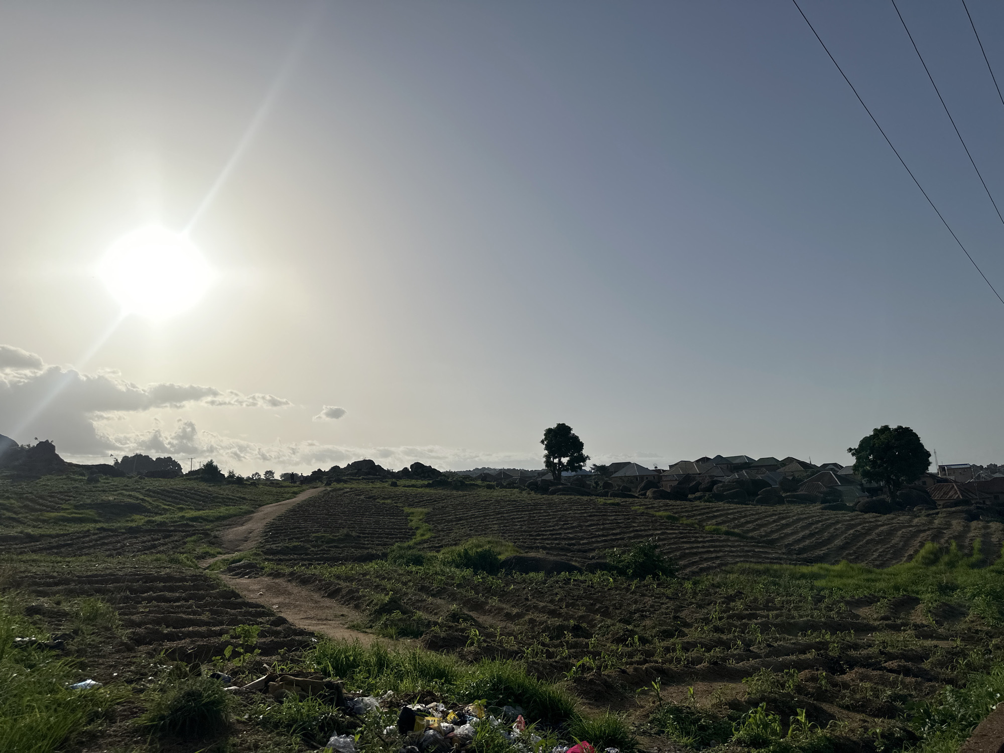 The image depicts a rural landscape in Jos, Nigeria, under a bright, hazy sun. The scene is dominated by terraced fields, likely cultivated with crops that are beginning to sprout, indicated by small green shoots. A dirt path winds through the fields, leading towards a cluster of houses and trees in the middle ground. The houses have corrugated iron roofs, a common feature in the region. In the foreground, there is a pile of scattered refuse.
The sun is high in the sky, casting a bright glare and creating a hazy atmosphere. The sky itself is a pale blue, with some wispy clouds. Electrical wires are visible in the upper right corner, indicating the presence of infrastructure. The overall impression is one of a rural agricultural setting, possibly early morning or late afternoon given the angle of the sun and the light quality. The presence of cultivation and homes suggests an inhabited area, while the discarded trash points to potential environmental concerns. No text is visible in the image. The scene evokes a sense of a working landscape, with a touch of everyday life and its challenges.