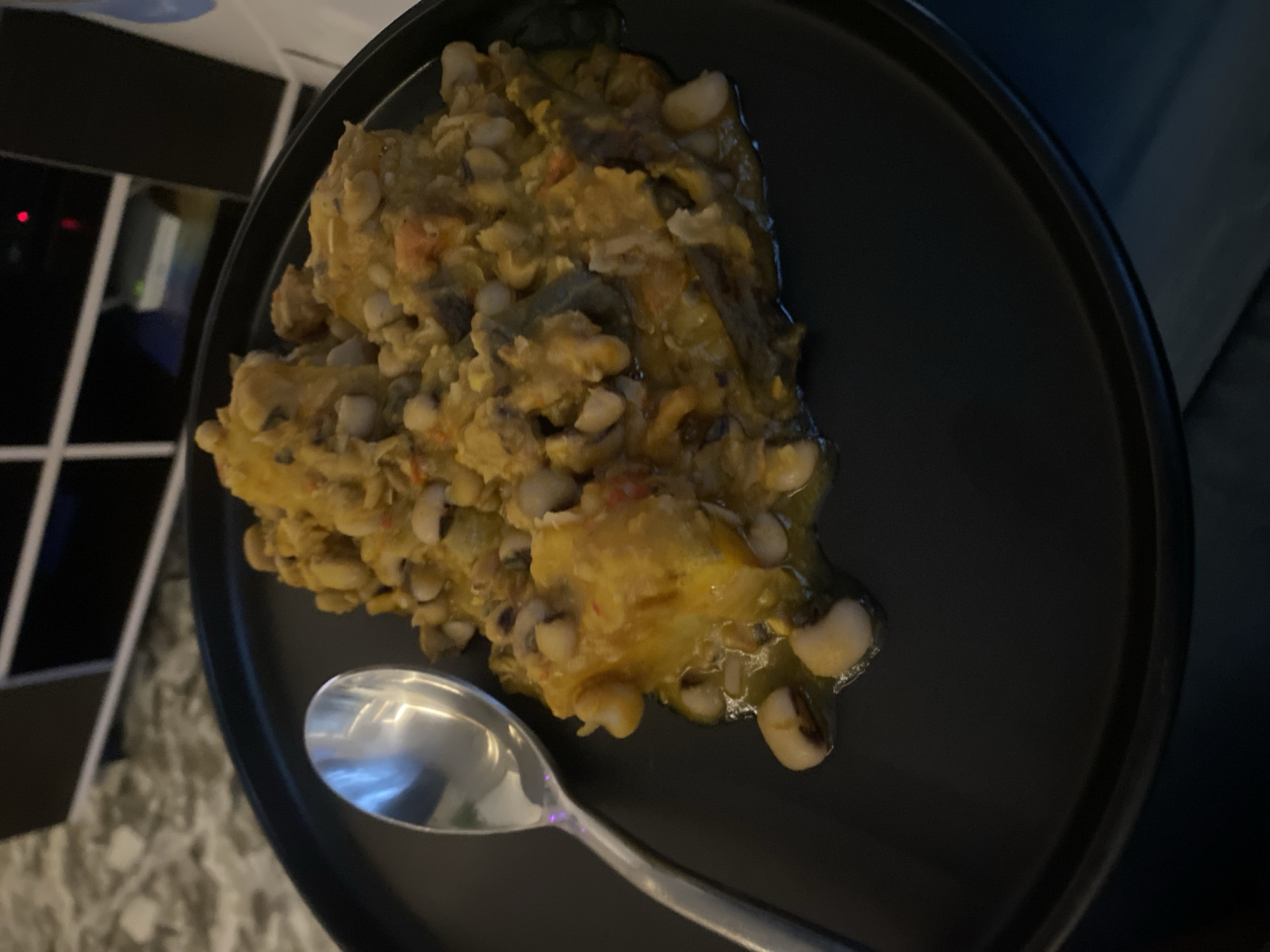 A plate of food, possibly black-eyed peas, is presented on a black plate alongside a spoon. The photograph appears to be taken indoors, possibly in Abuja, Nigeria.