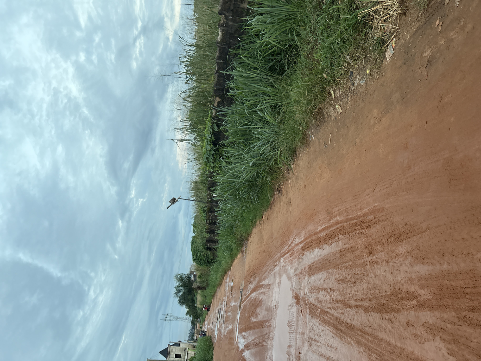 A dirt road in Obosi, Nigeria, is shown from a low angle, looking down the path. The road is muddy with visible tire tracks and puddles, indicating recent rain. To the left, a partially constructed building with a dark, pointed roof and a tall electricity pylon are visible. Two motorcycles are parked near the building. Tall green grass and dense vegetation line the right side of the road, forming a natural barrier along a low, dark stone wall. The sky above is overcast with dramatic, textured clouds. The scene conveys a sense of a rural or developing area with rough terrain.